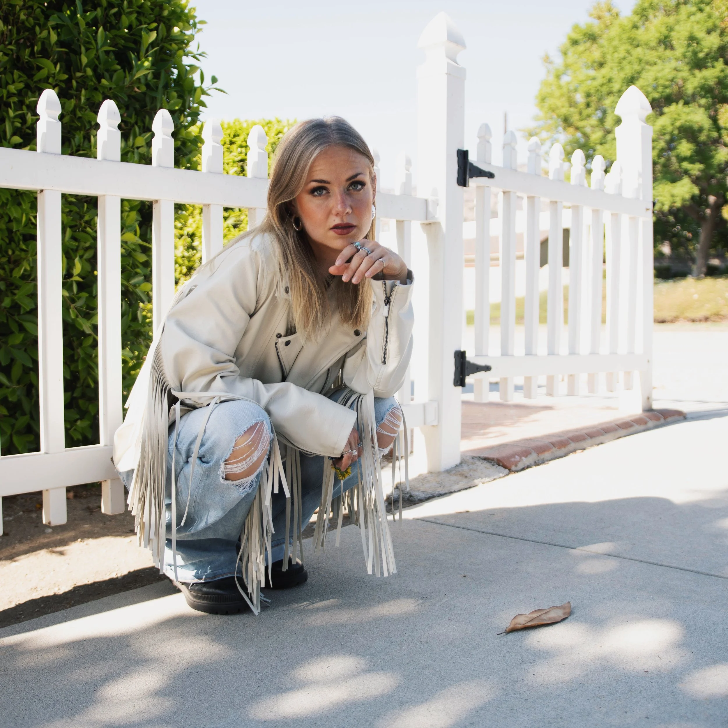 A young woman with blonde hair, wearing a white jacket and ripped jeans, squats by a white picket fence on a sidewalk. She has multiple rings and earrings, and is looking directly at the camera with a serious expression. A fallen leaf is on the ground, and green bushes and trees are in the background under clear skies.
