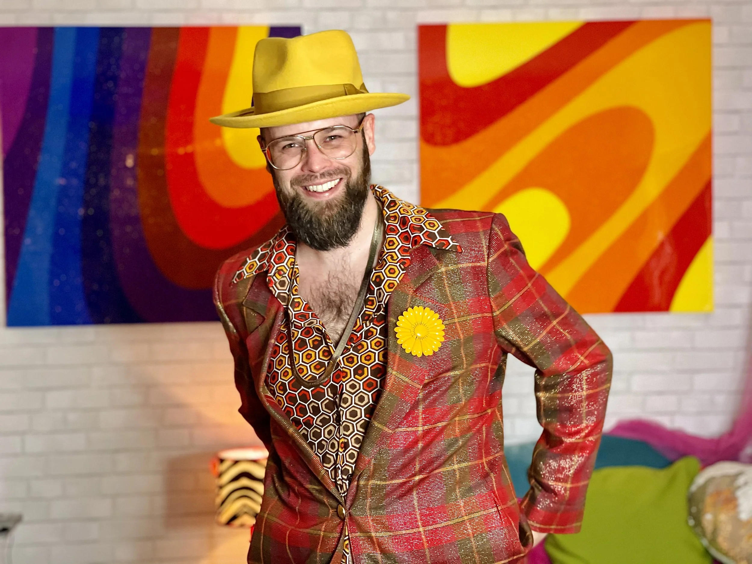 Artist and DJ Harald Austad wearing a yellow hat and a colorful, patterned blazer with a yellow flower pin, standing in front of a vibrant abstract painting.