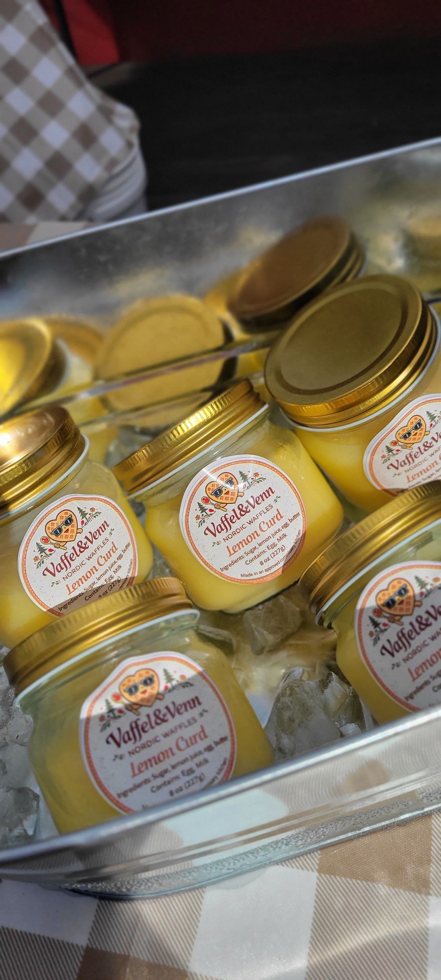 Glass jars of lemon curd with gold lids and Christmas-themed labels, placed in a metal container with ice, on a checkered tablecloth.