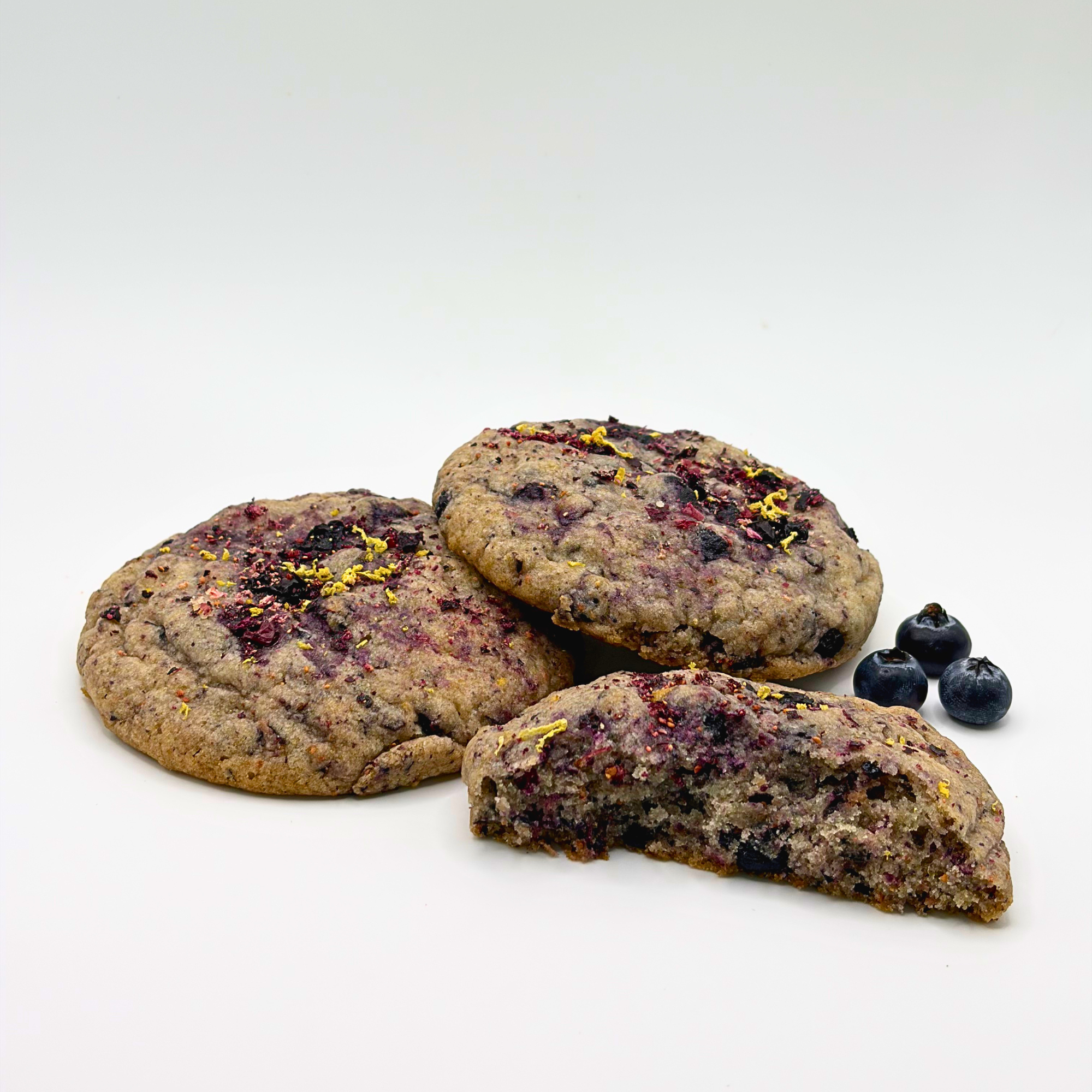 Three oatmeal cookies decorated with dried berries, slices of dried lemon, and colorful flower petals, arranged on a white surface with blueberries nearby and a plain white background.