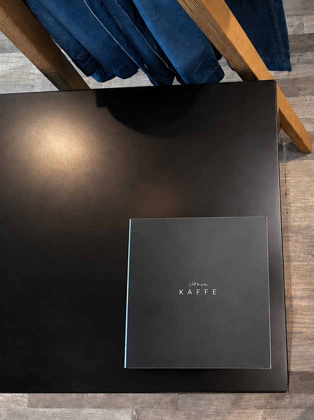 Black table with a brand book labeled 'KAFFE' in the centre at Susan Cooper Agency for wholesale fashion in Vancouver, BC. In the background, a clothing rack with folded denim jeans hanging on it.