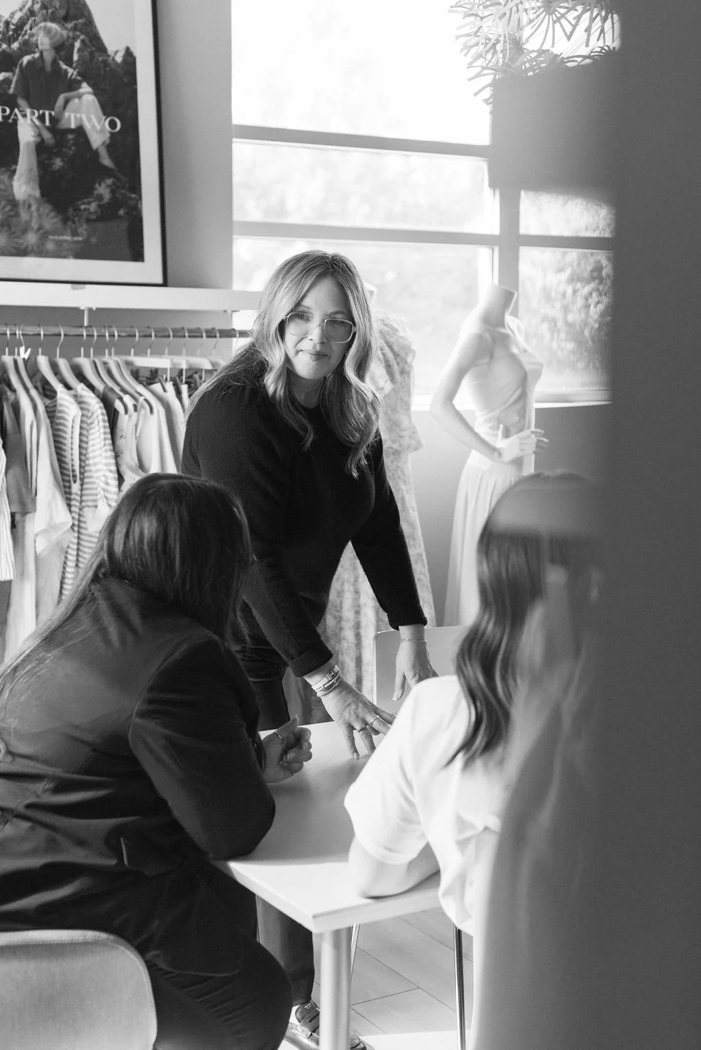 A woman, Susan Cooper, with glasses standing at the showroom table, engaged in discussion with the Susan Cooper Agency sales team, with clothing racks and a mannequin in the background.