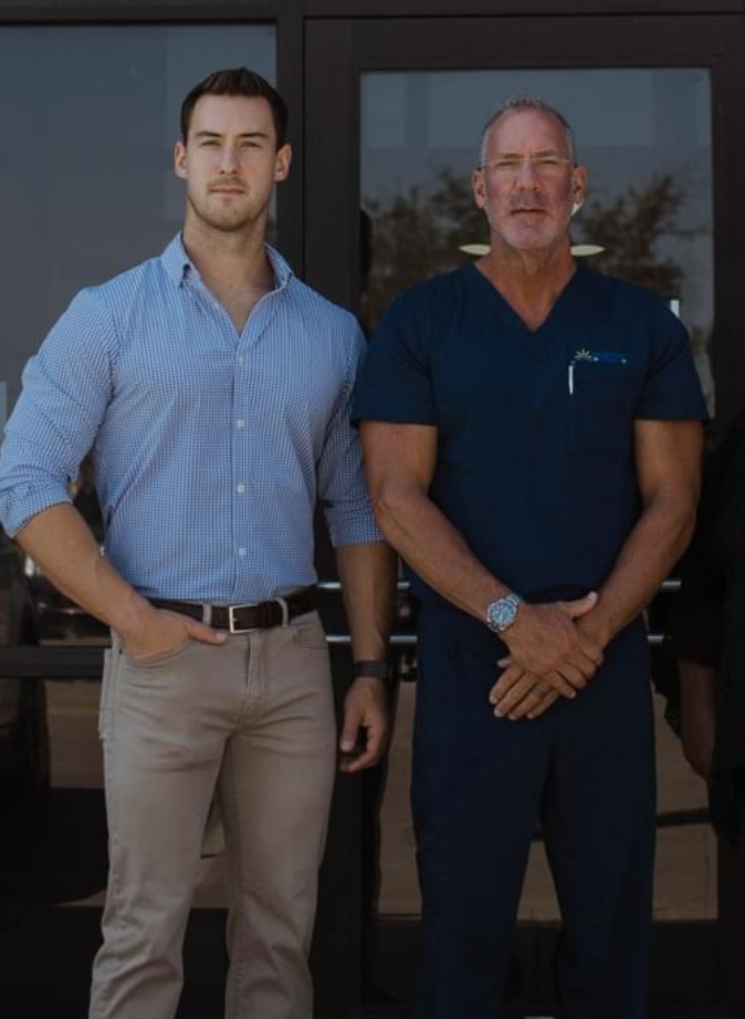Two men standing outside against a dark-framed glass door, one young with dark hair wearing a light blue checkered shirt and beige pants, the other older with gray hair and glasses wearing navy scrubs and a watch.
