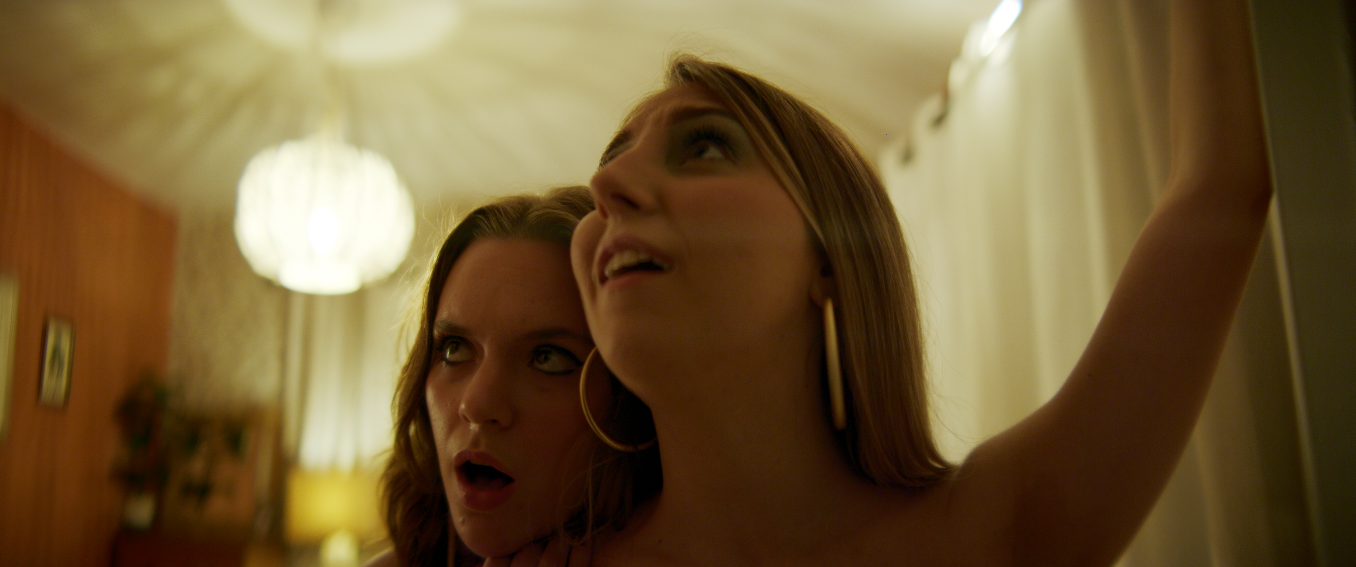 Two women in a room with warm lighting, one of them holding a selfie stick, both looking up with expressive faces.