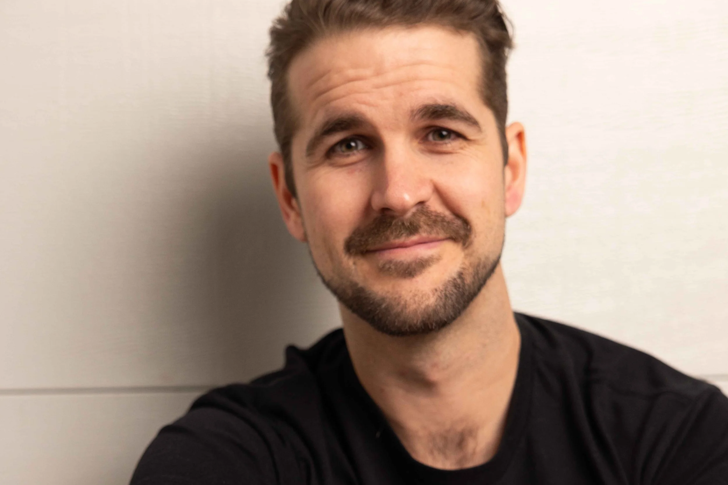 A man with light skin, short brown hair, a beard, and light-colored eyes smiling and looking at the camera, wearing a black shirt, against a plain light-colored background.