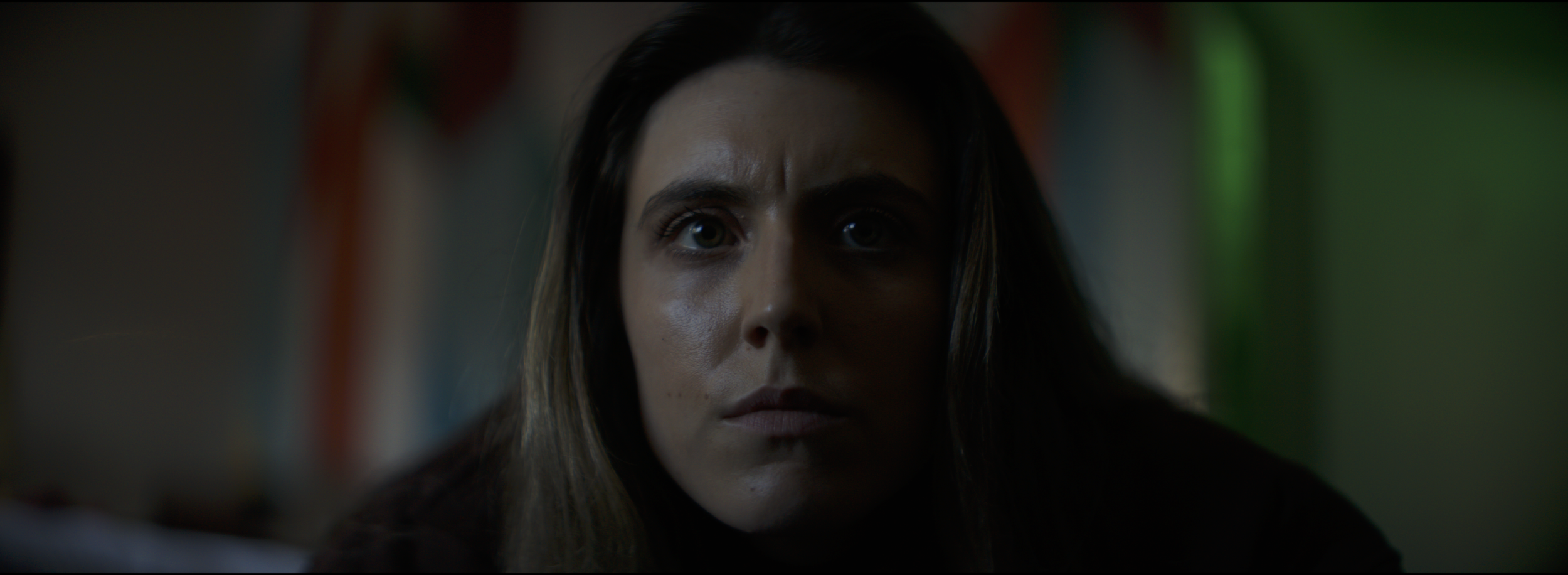 Close-up of a woman's face with intense expression, partially shadowed, in a dark environment with colorful blurred background lights.