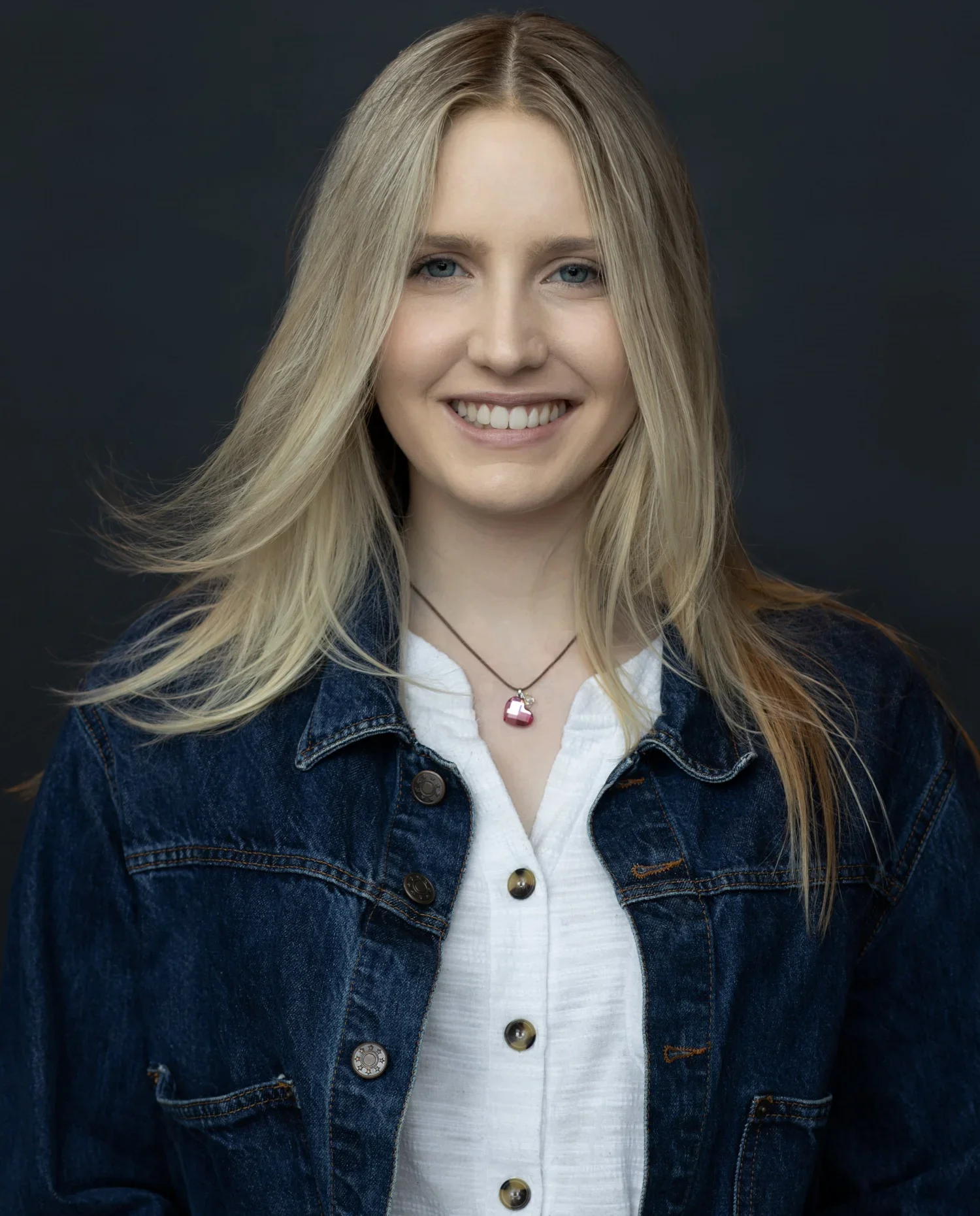 Close-up portrait of a young woman with long blonde hair, blue eyes, and a bright smile, wearing a denim jacket over a white shirt and a pink pendant necklace, set against a dark background.