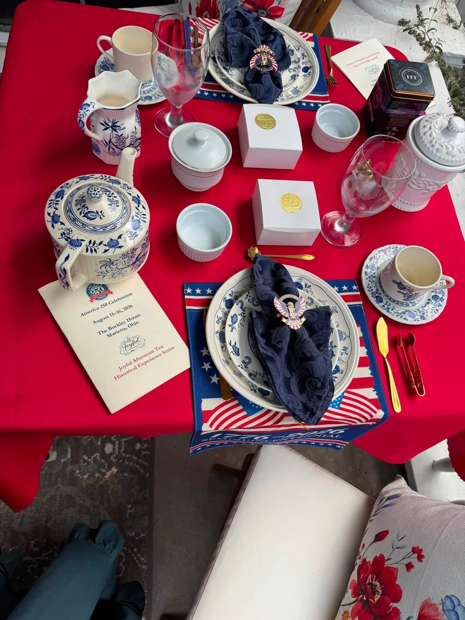 A table decorated for a patriotic celebration with red, white, and blue themed tableware, including plates, cups, and napkins. There are teacups, wine glasses, boxes, and a teapot, alongside a small booklet or invitation and a decorative medal. The tablecloth is red, and part of a floral-patterned cloth and a white bench are visible.