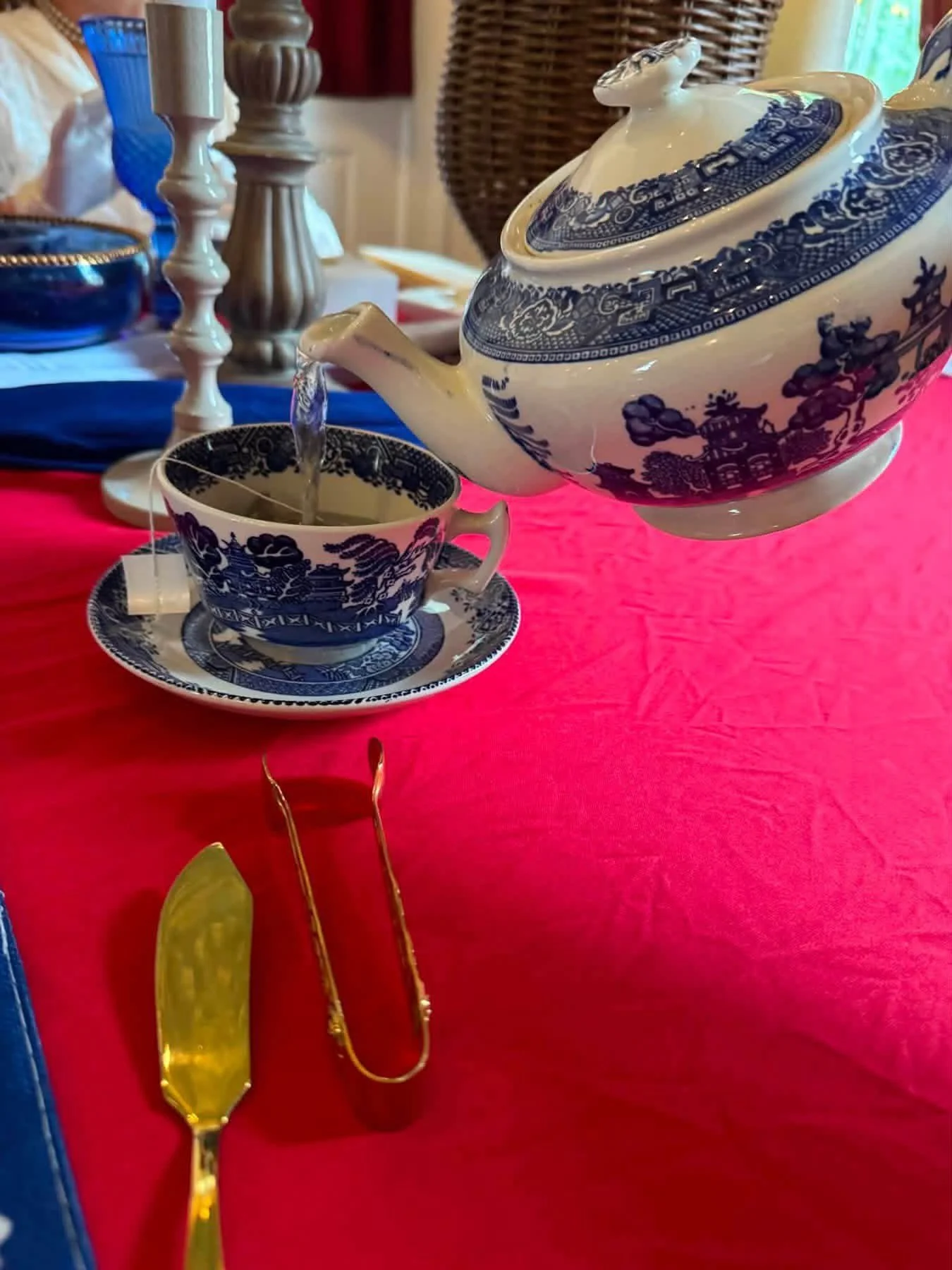 A teapot pouring tea into a matching teacup and saucer on a pink tablecloth, with a yellow serving utensil and pair of gold-colored tongs in front of the table.