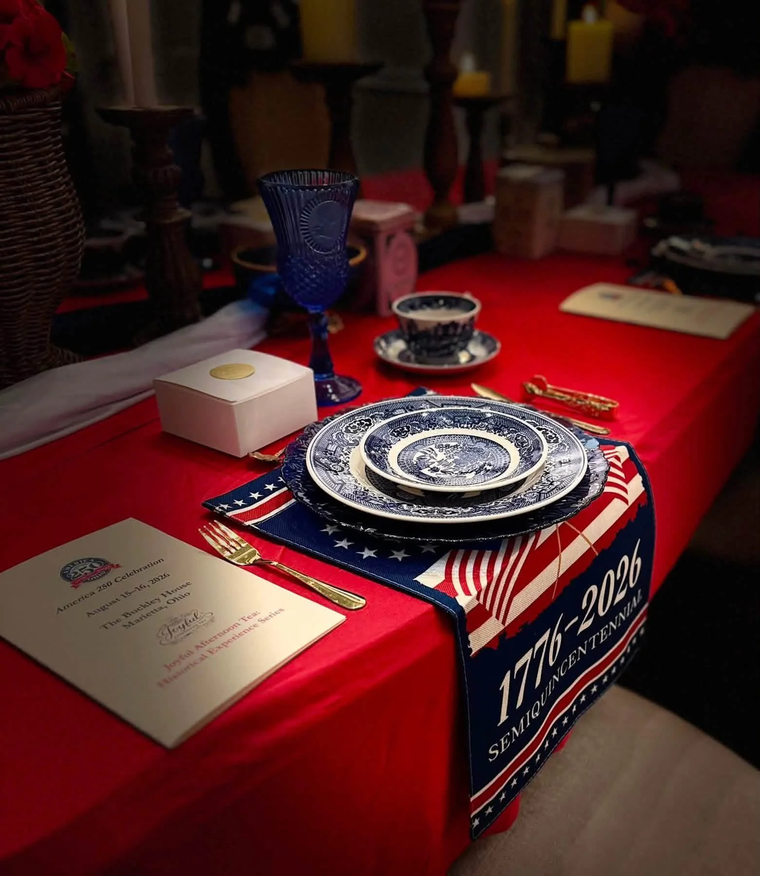 Table with red tablecloth set for a commemorative event, featuring blue and white patterned plates, a blue glass, a teacup and saucer, gold utensils, and a program for the America 250 celebration.