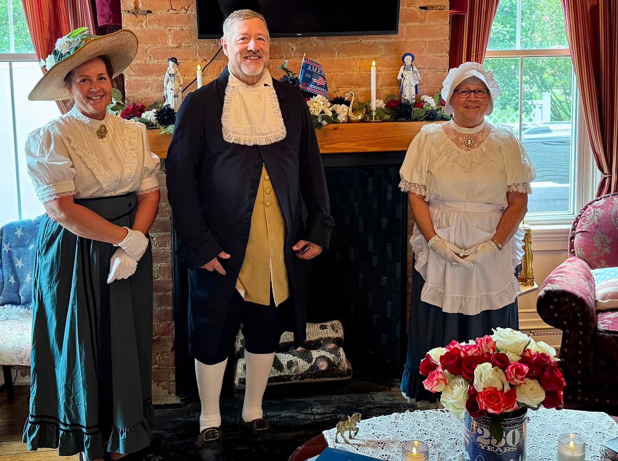 Three people dressed in historical clothing standing indoors in front of a brick fireplace decorated for a celebration, with a bouquet of roses and a candle on the table in front.