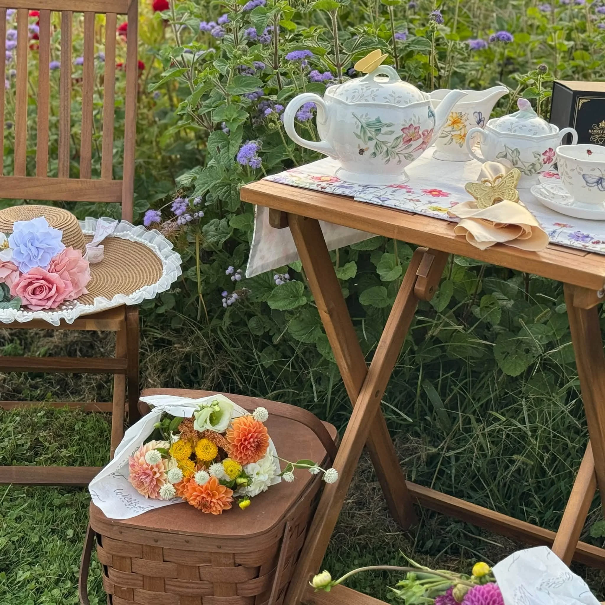 Tea set with floral patterns on a wooden table outdoors, surrounded by purple flowers and green foliage.