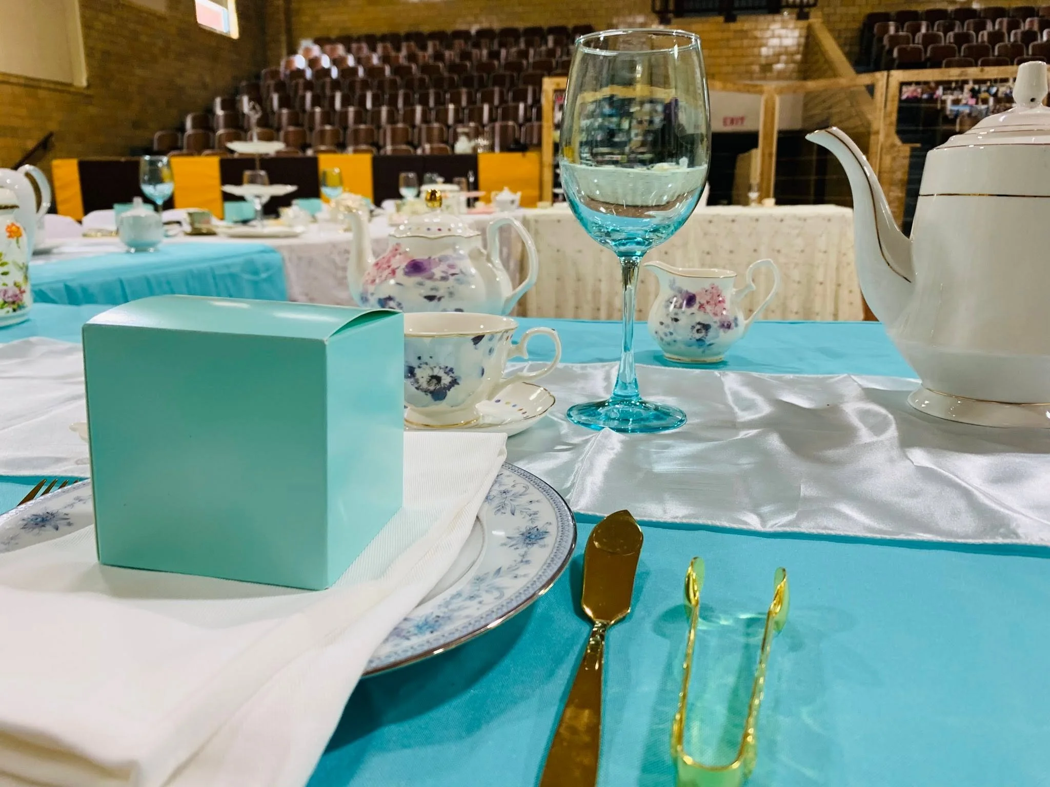 A table set for a formal event with china, a glass of water, a teapot, and a Variety Tea Sachet box on a turquoise tablecloth. In the background, there are more tables and empty chairs.