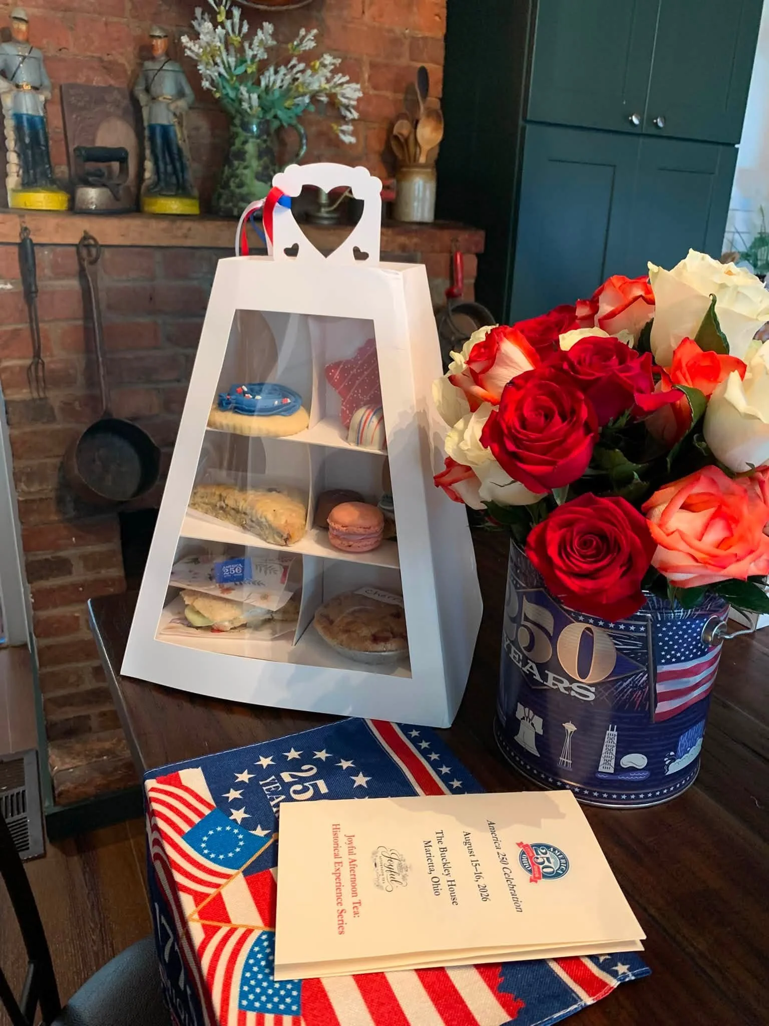 Celebration table featuring a certificate of appreciation, a bouquet of red, white, and peach roses, a white gift bag with heart-shaped cutouts, a small decorative wreath, and a patriotic-themed tablecloth. The background includes a brick wall with vintage lanterns and a blue cabinet.