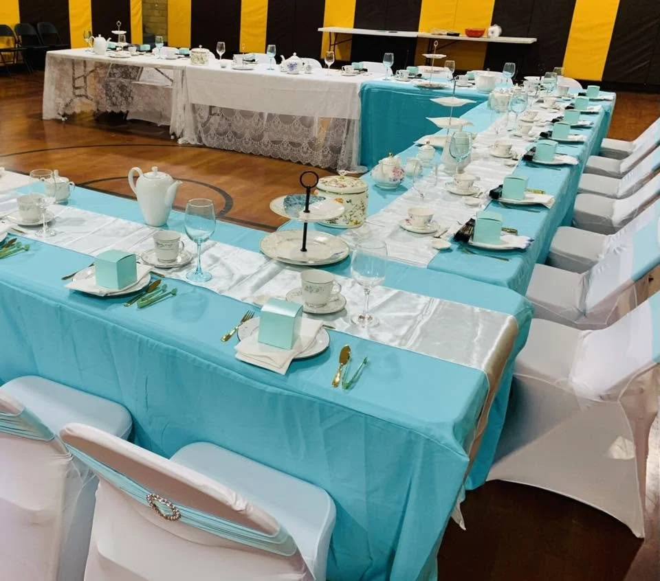 A long dining table set for a formal event with turquoise tablecloth, white table runner, and white chairs. The table features tea sets, glassware, small boxes, and decorative accessories, with additional tables in the background covered with lace tablecloths.