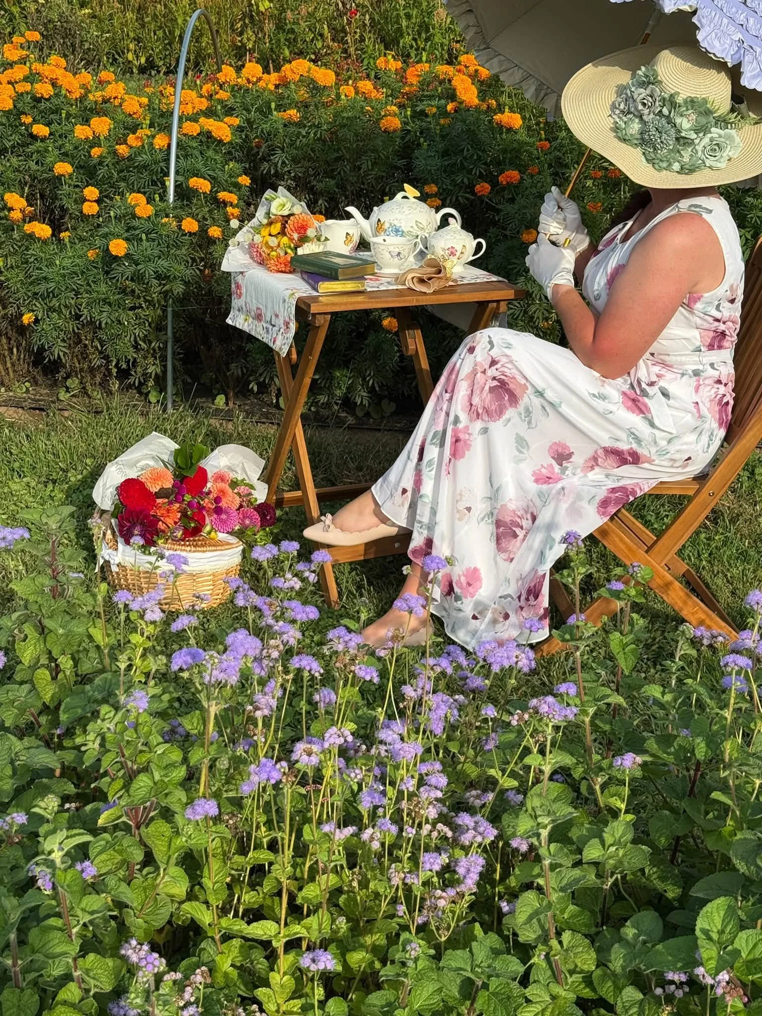 A woman in a floral dress with a large sun hat sitting on a wooden chair in a garden with orange marigolds and purple asters, with a table of tea items and flowers in front of her.