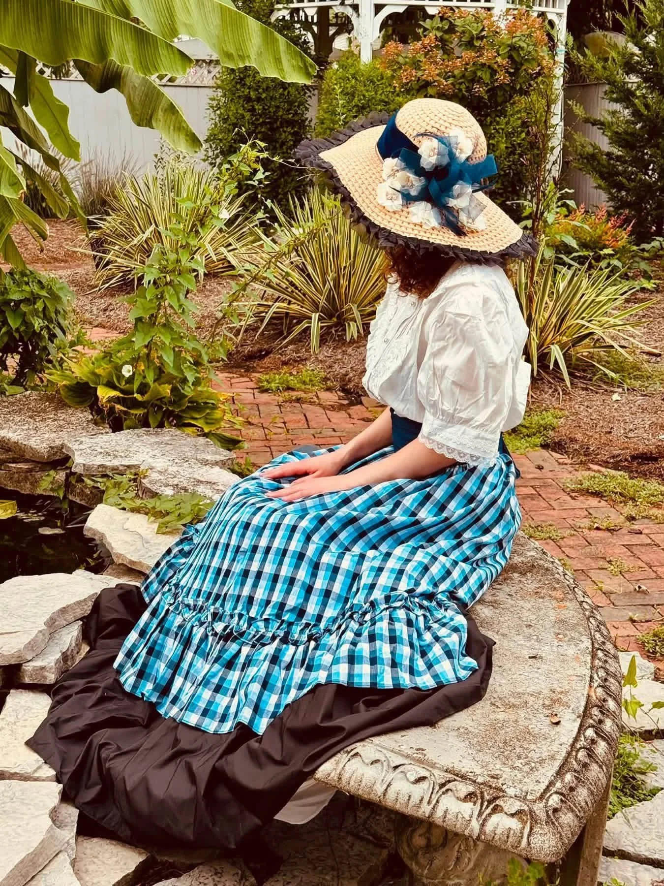 A woman wearing a large sun hat with a blue ribbon, a white blouse, and a blue plaid skirt sitting on a stone bench in a garden with various green plants and shrubs.