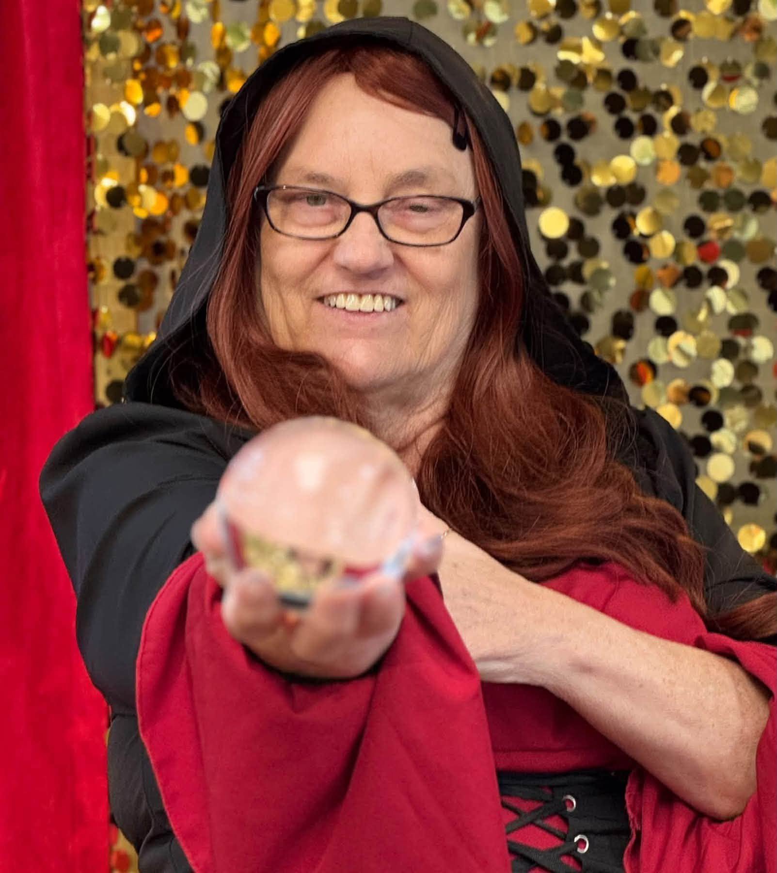 A person with long red hair, glasses, and a black hooded jacket is smiling and extending their hand forward, appearing to offer something. The background features a sparkling gold sequin backdrop and a red curtain on the left side.
