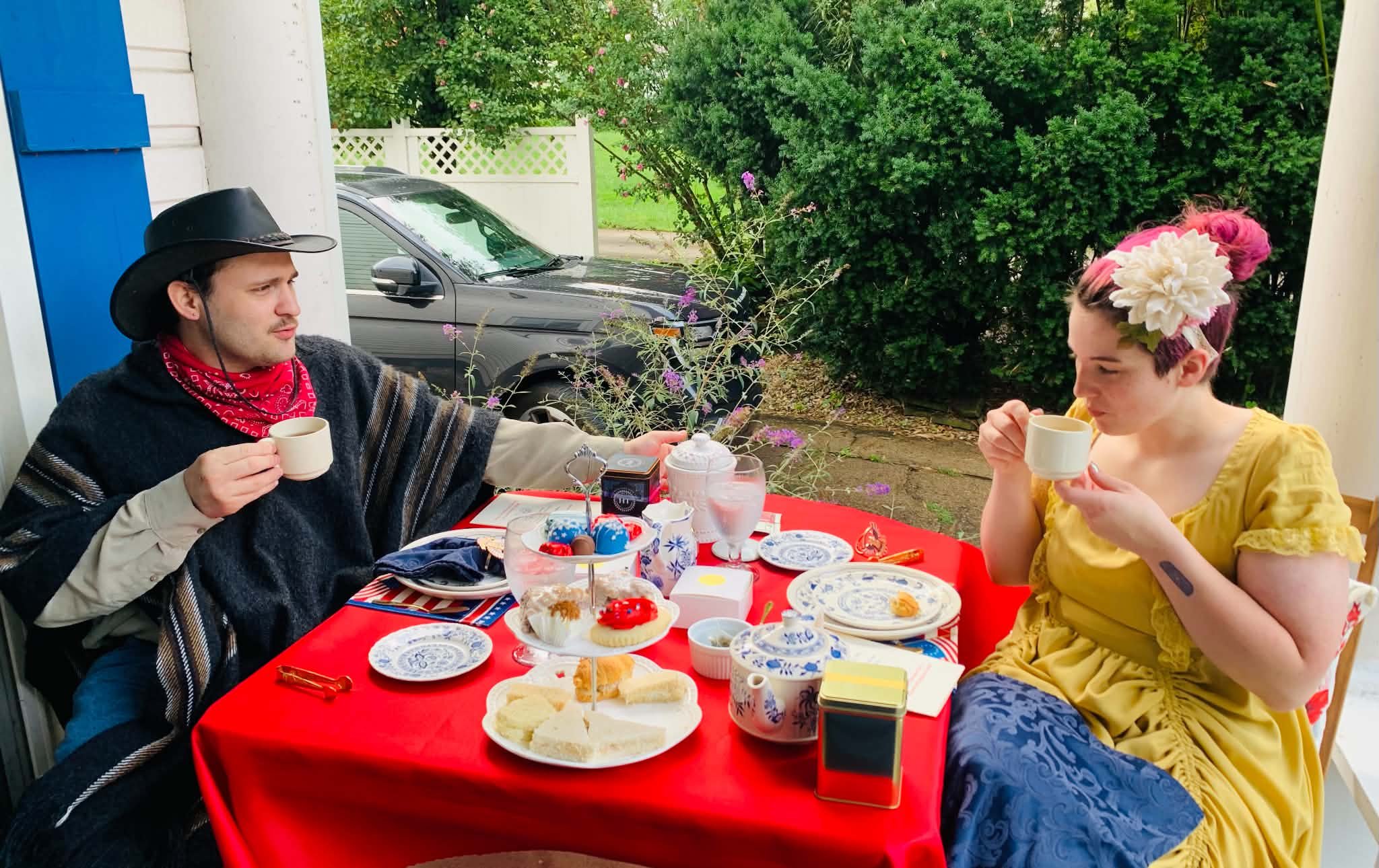 A young man dressed as a cowboy and a young woman with a floral headpiece sit at a table outdoors, enjoying tea and desserts on a sunny day with greenery and a black car in the background.