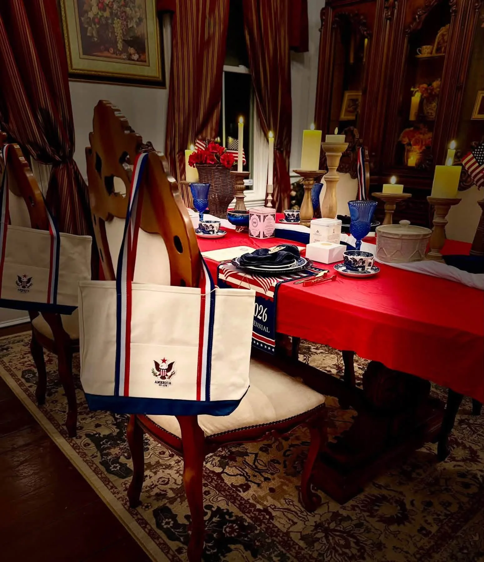 A dining table decorated for a patriotic American celebration with red, white, and blue colors, American flags, candles, and patriotic themed tableware. The table is surrounded by wooden chairs, and the background features a wall with a painting, curtains, and a wooden cabinet with decorative items.