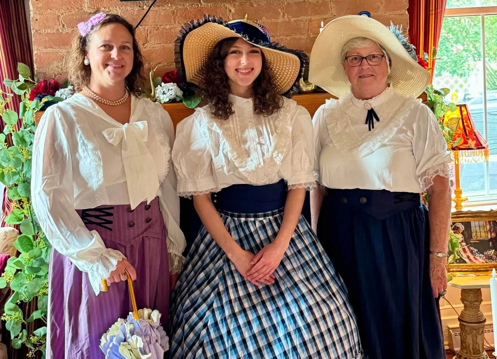 Three women in vintage-style clothing and large hats standing indoors in front of a brick wall with flowers, greenery, and a window in the background.