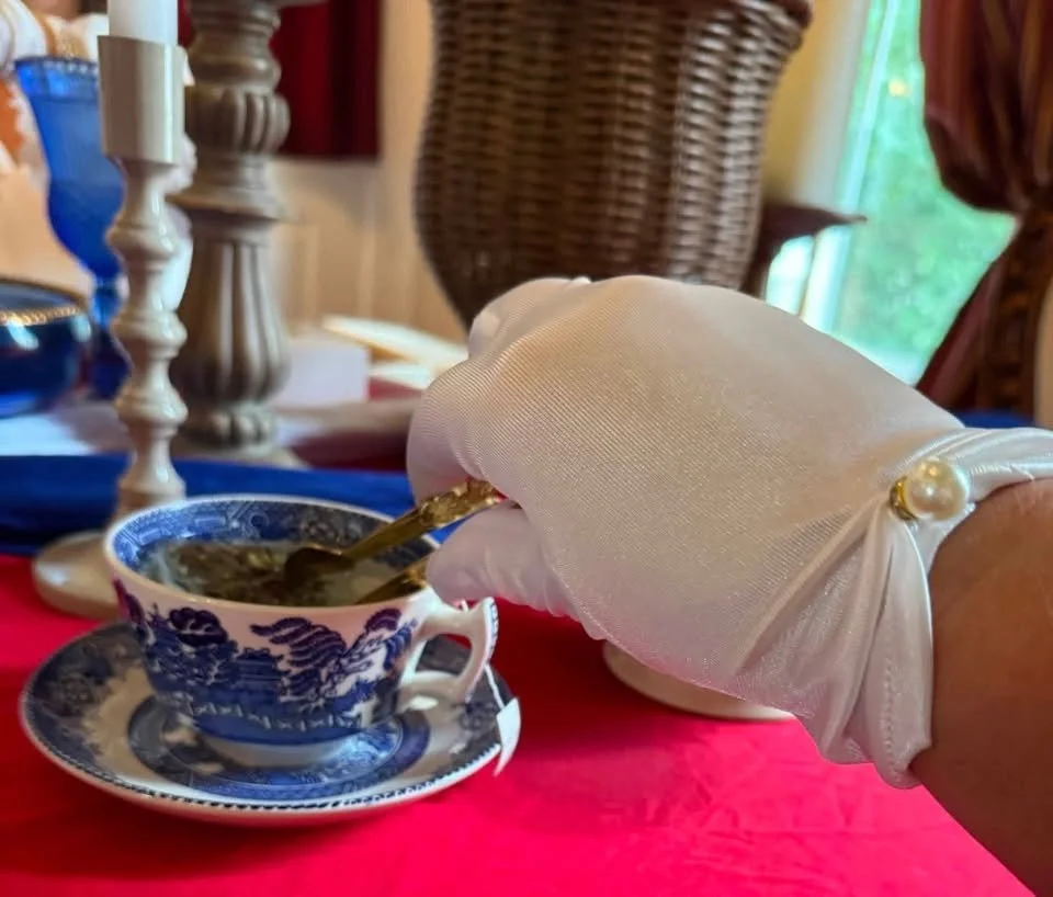 Person wearing white gloves holding a spoon and scooping jam or jelly from a decorated tea cup with a matching saucer, on a red tablecloth, with candles and a wicker basket in the background.