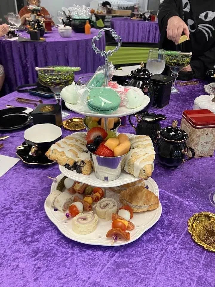 A three-tiered dessert stand on a purple tablecloth, featuring assorted pastries on the bottom, fruit and small desserts on the middle, and decorated cakes on the top, with teapots, glasses, and other tableware nearby.