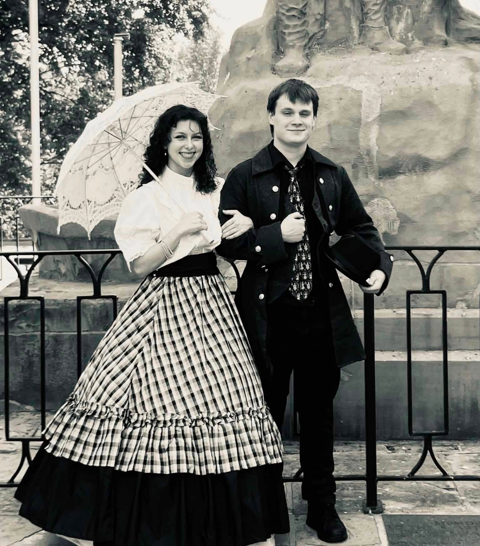 A black-and-white photo of a young woman and young man at a park, standing in front of a fence and a large rock sculpture. The woman is smiling, holding a lace umbrella, and wearing a white blouse and a long checkered skirt. The man has a slight smile, with his arms crossed, and is dressed in a dark jacket, shirt, and tie.