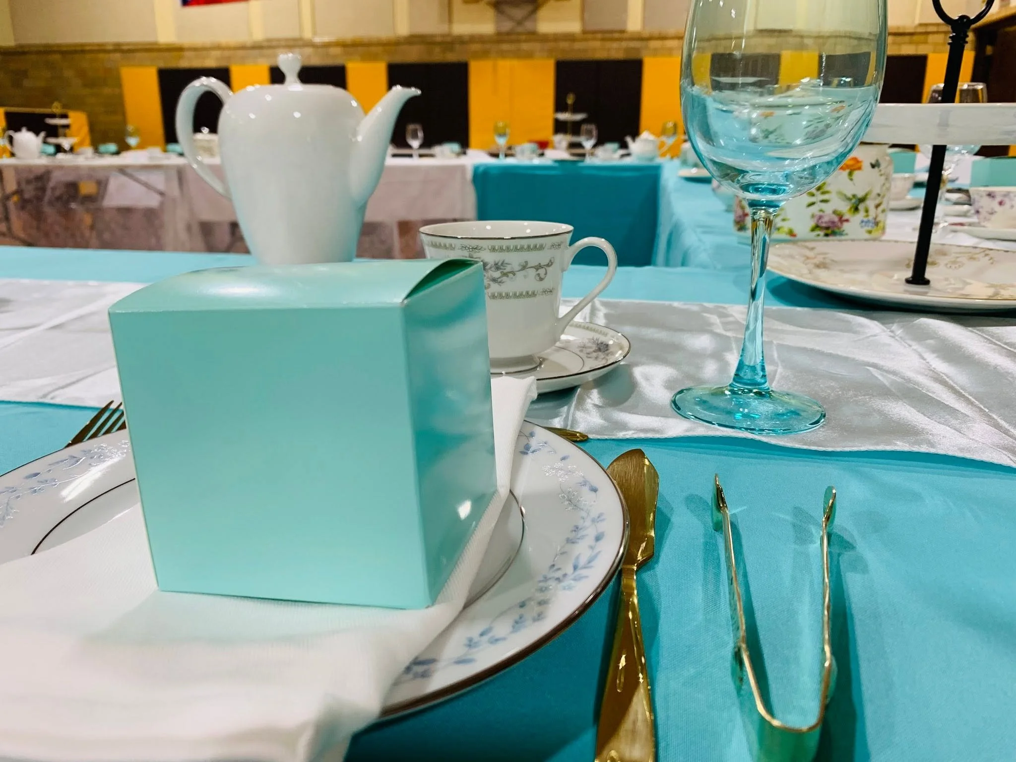 A table set for a formal event with white and teal tablecloths, fine china, glassware, and gold utensils. A white teapot, a glass of water, a teacup with a saucer, and a gift box with a lid are visible in the foreground. The background shows more tables and yellow and black padded wall panels.