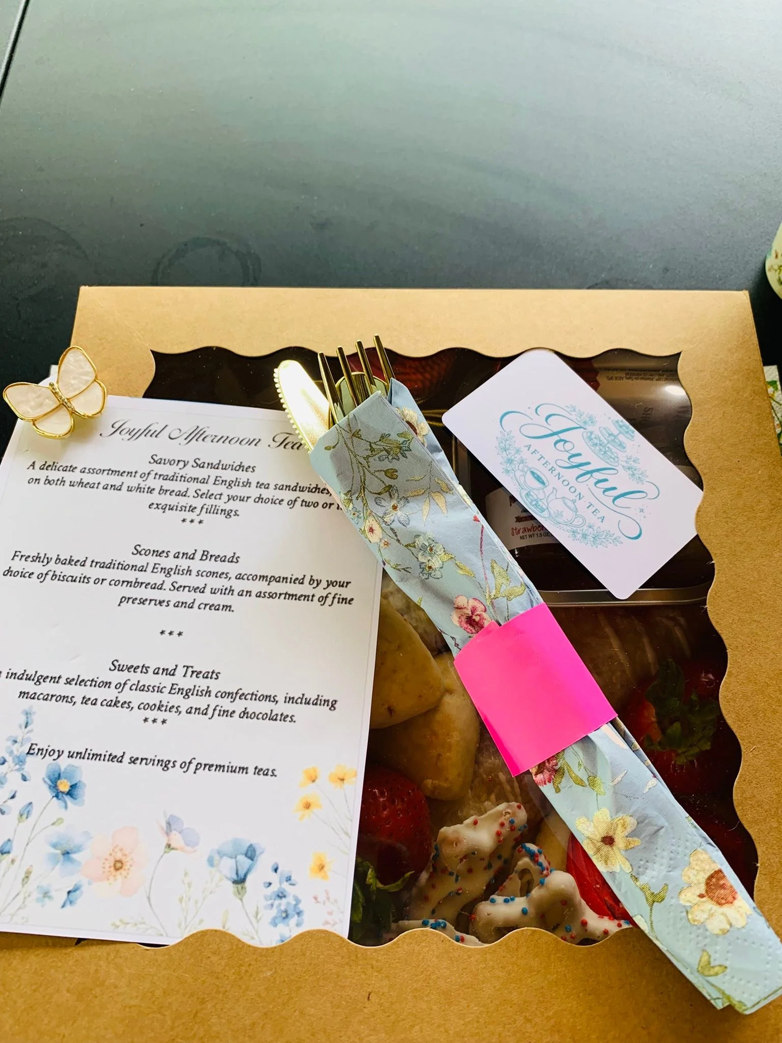 A partially opened bakery box containing assorted cookies, with a floral-themed paper napkin, a welcome card, and a menu for afternoon tea.