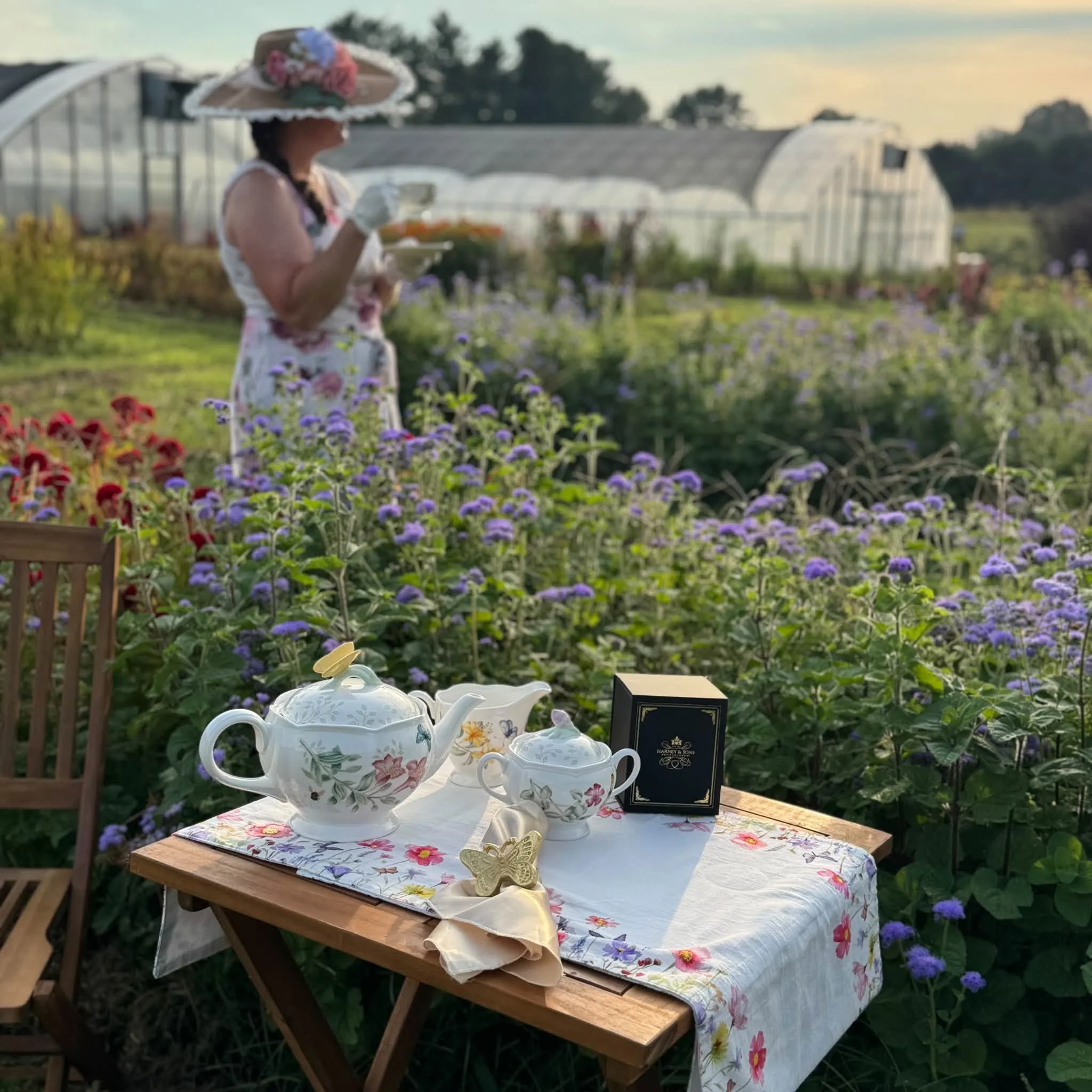 Table set for tea with white porcelain teapot, cups, and saucers on a tablecloth with floral pattern, outdoors among purple flowers with greenhouses and trees in the background at sunset.