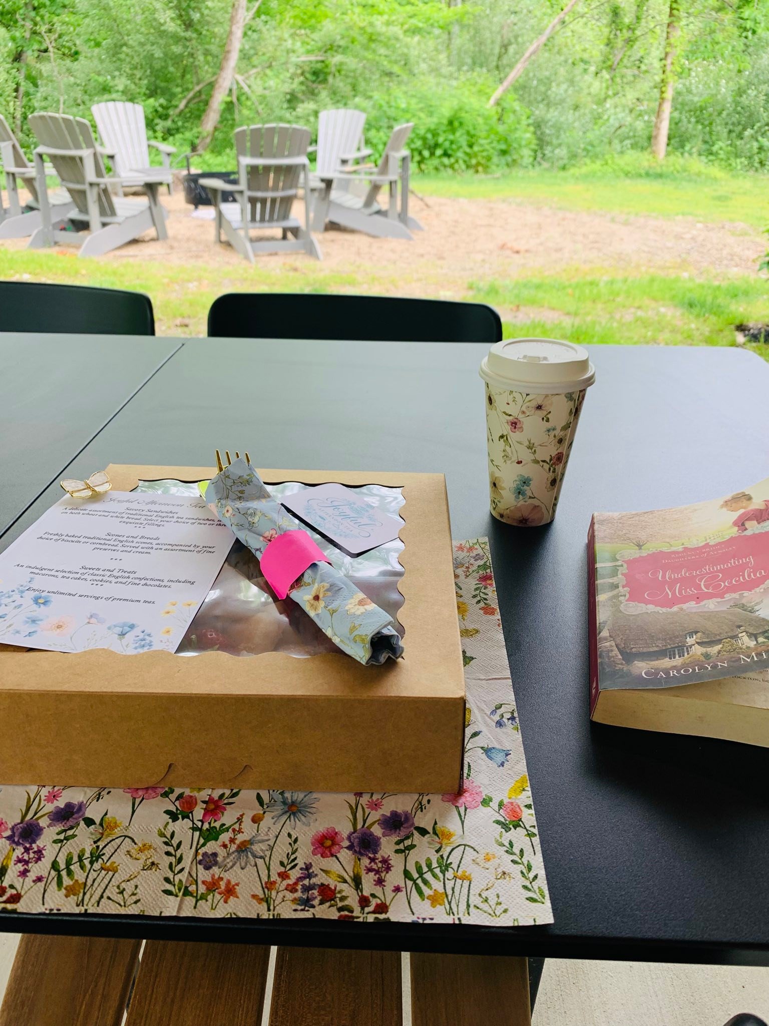 Table with a floral tablecloth, a takeaway coffee cup with a floral design, a menu, a notepad, a paper with writing, reading glasses, a wrapped pastry, and a book titled "Understanding Miss Cecilia" by Carolyn M. in an outdoor setting with green trees and Adirondack chairs in the background.