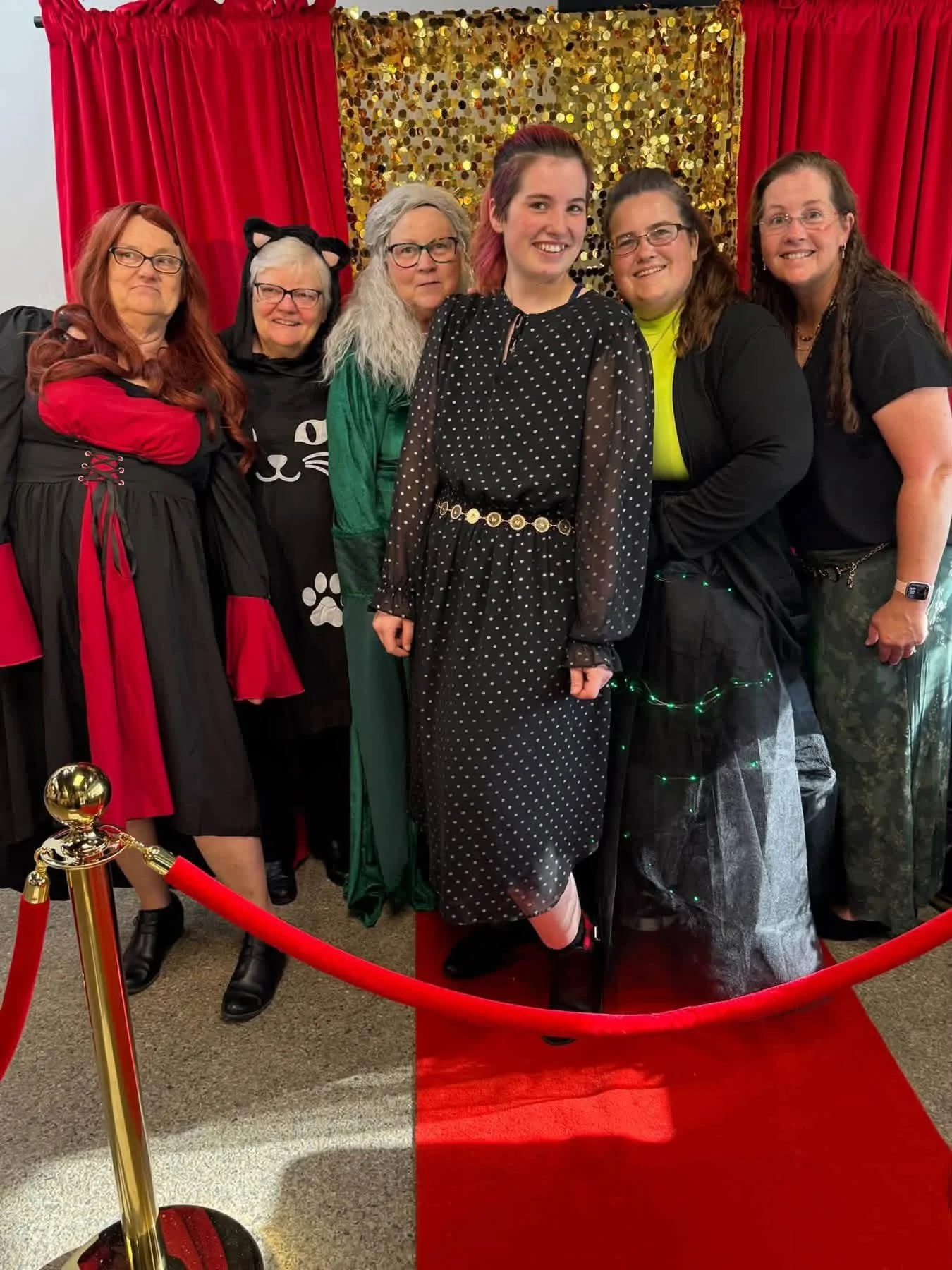 Group of six people dressed in costumes standing behind red velvet rope barrier on a red carpet, with a gold sequin backdrop and red curtains in the background.