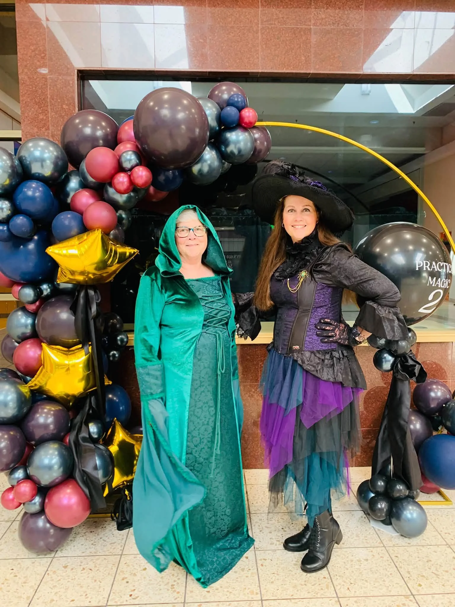 Two women dressed in elaborate costumes standing in front of a balloon arch with black, gray, blue, pink, and gold balloons, and a black balloon with gold writing that says 'PRACTICAL MAGIC'.