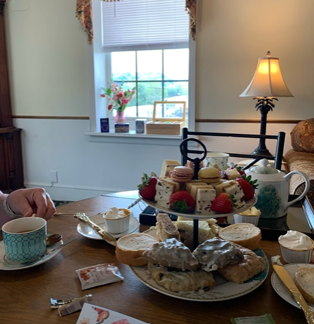 A table set for tea with various desserts, scones, cakes, strawberries, and tea cups. In the background, a window with a vase of pink flowers on the windowsill and a lamp on a side table.
