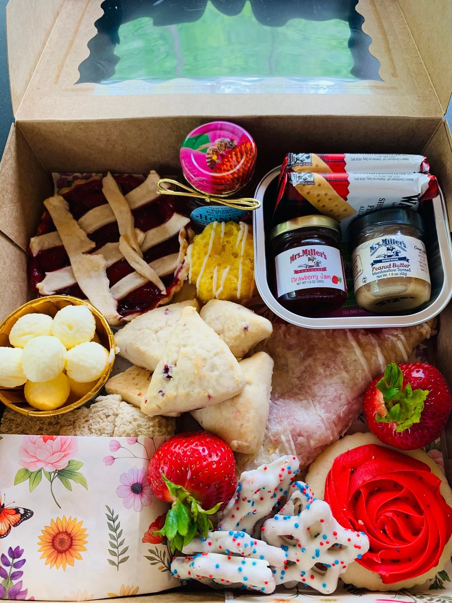A cardboard box with clear window containing assorted baked goods, jams, cookies, strawberries, and whipped cream.