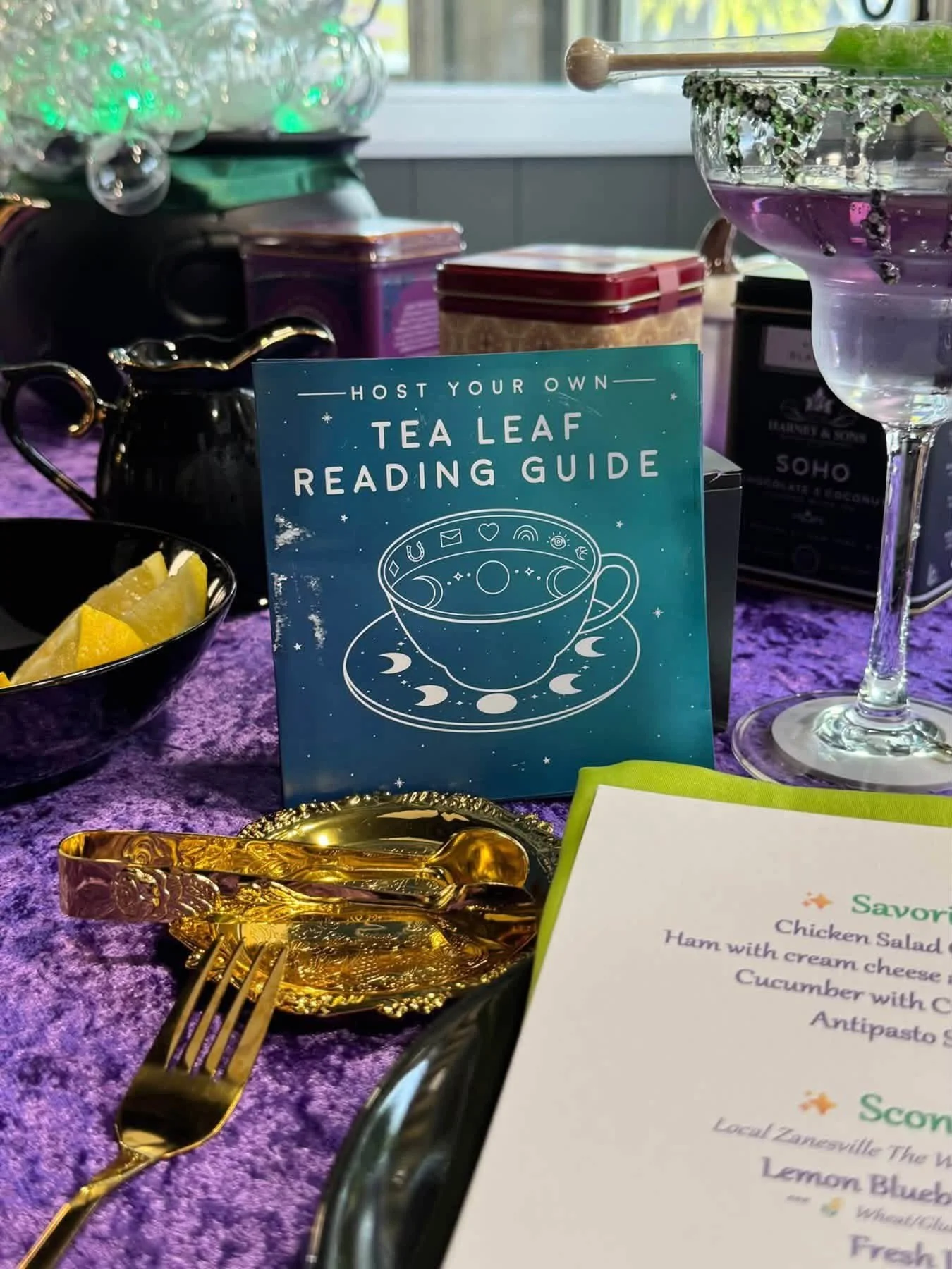 A table setting with a teal sign that reads 'Host Your Own Tea Leaf Reading Guide,' a gold-colored fork and spoon, a black cup with lemon wedges, a menu with food items, and decorative glassware on a purple tablecloth.