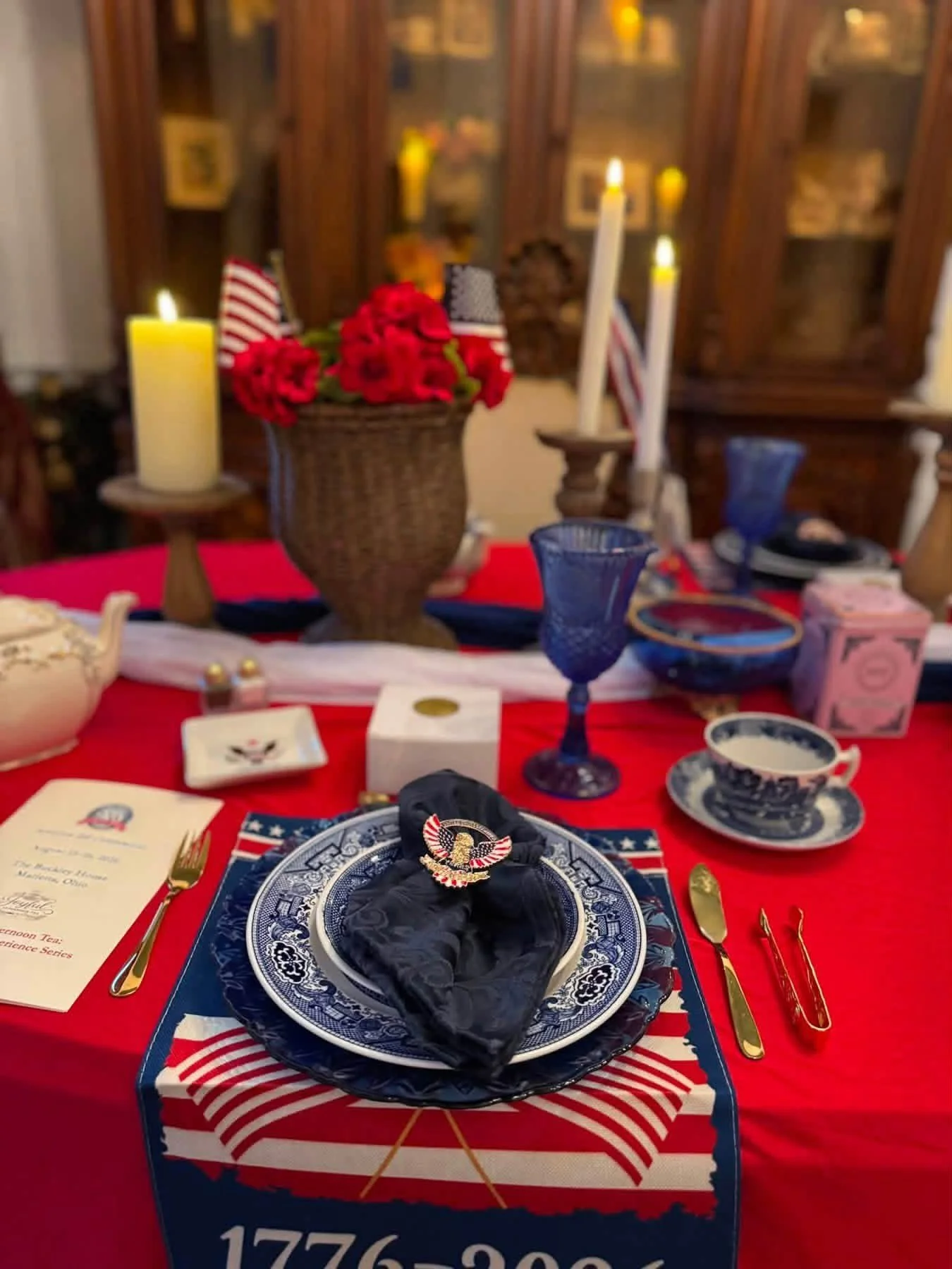 A table set with patriotic-themed decorations, including red, white, and blue plates, gold utensils, blue glasses, and a black napkin with a patriotic badge. The background features candles, flowers, and American flags, with a wooden cabinet displaying framed photographs or artwork.