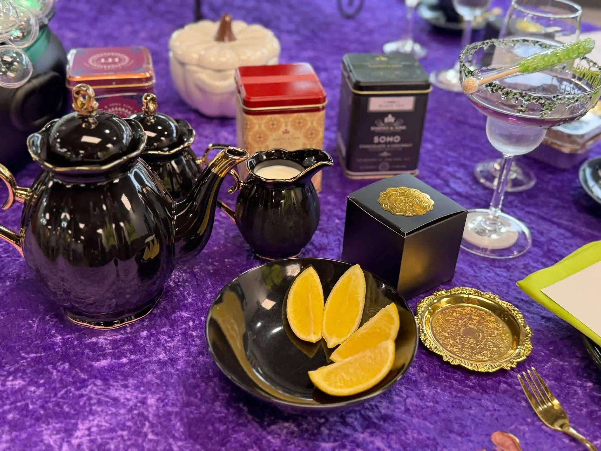 Black teapot and creamer set on purple tablecloth, alongside small black bowls with lemon wedges, decorative tins,  and drinking glasses.