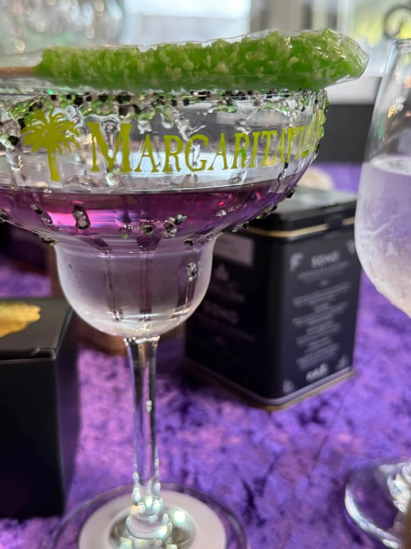 A margarita cocktail in a branded glass with a lime wedge on top, filled with a pinkish-purple drink and some ice, sitting on a purple textured surface.