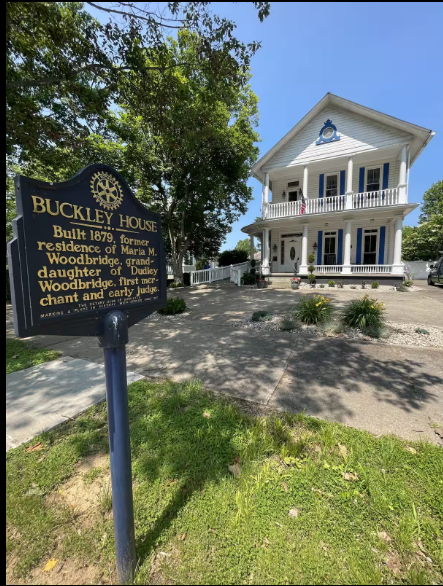 A historic white wooden house with a porch and balcony, next to a sign indicating it is Buckley House, built in 1879, and once the residence of Maria M. Woodbridge.