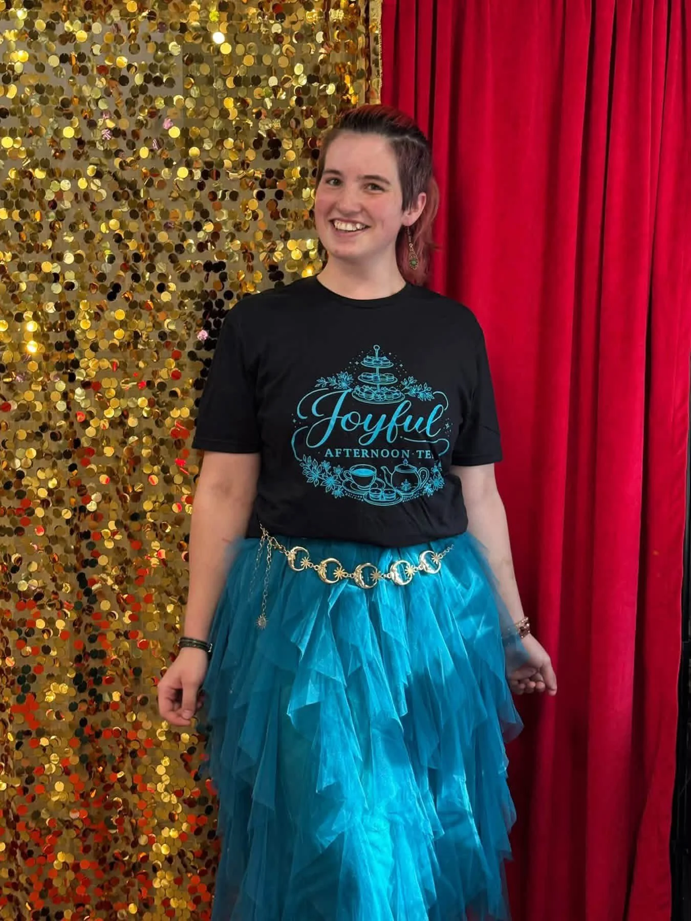 A smiling young woman wearing a black t-shirt with the words 'Joyful Afternoon Tea' and a teapot graphic, paired with a blue tulle skirt, stands in front of a gold sequin backdrop and red velvet curtain.