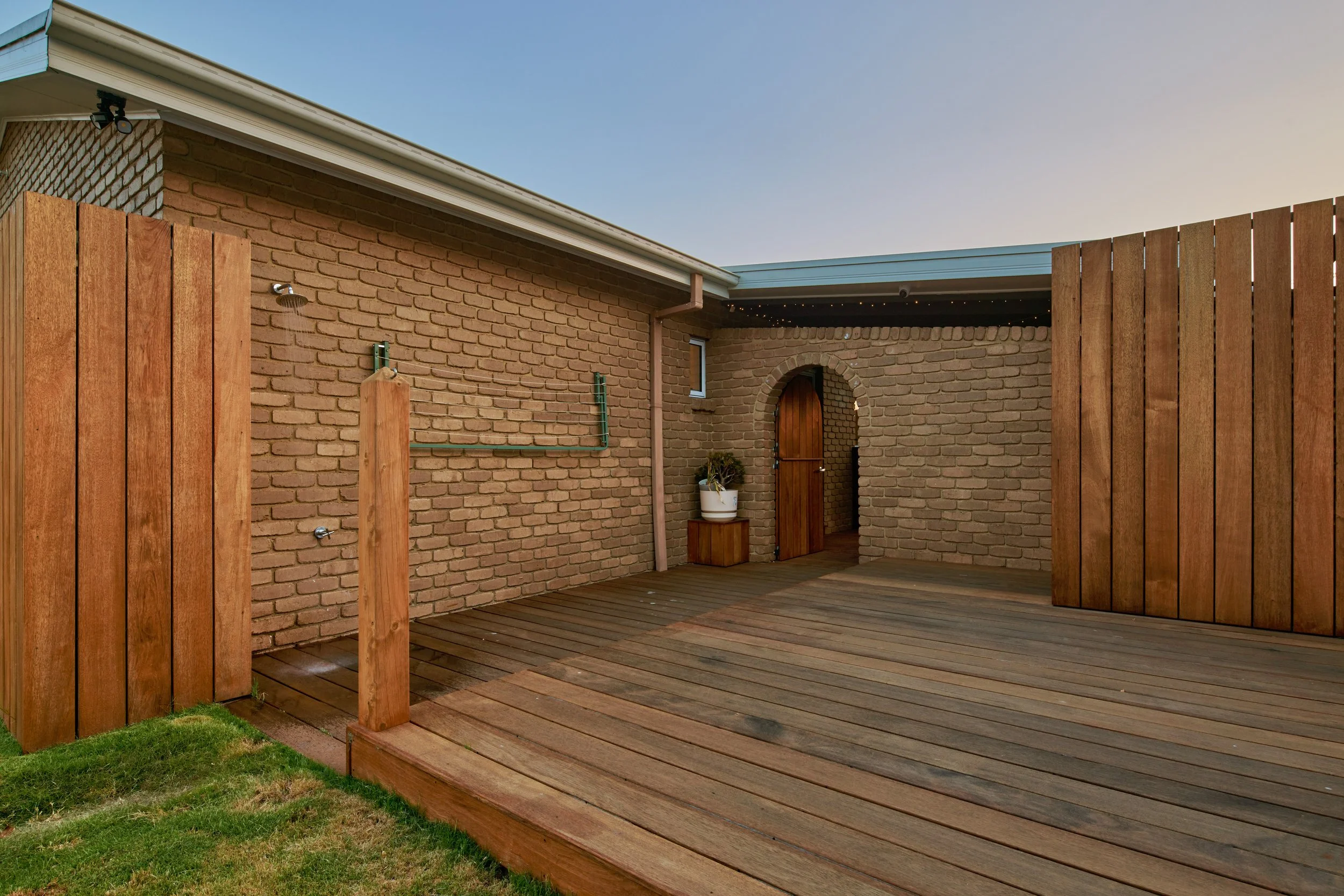 Backyard patio area with wooden deck, brick wall, and a small arched wooden door, with fencing and a potted plant.