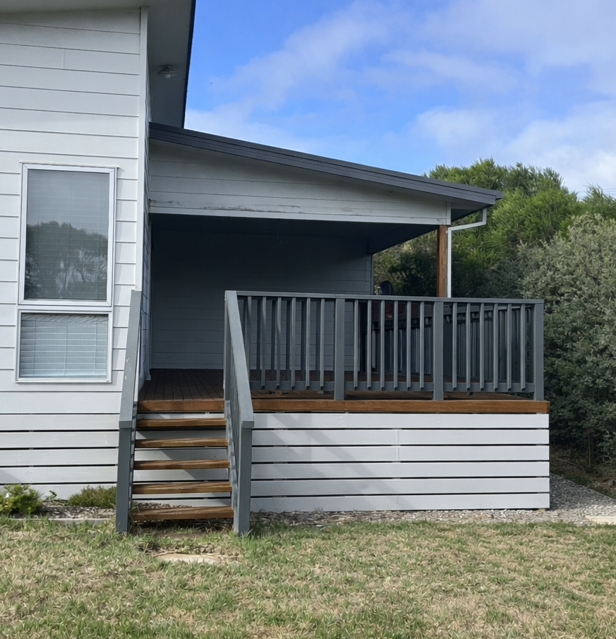 A white house with a small balcony featuring a wooden floor and gray railing, accessible by a short staircase, with a lawn in the foreground and trees in the background under a partly cloudy sky.