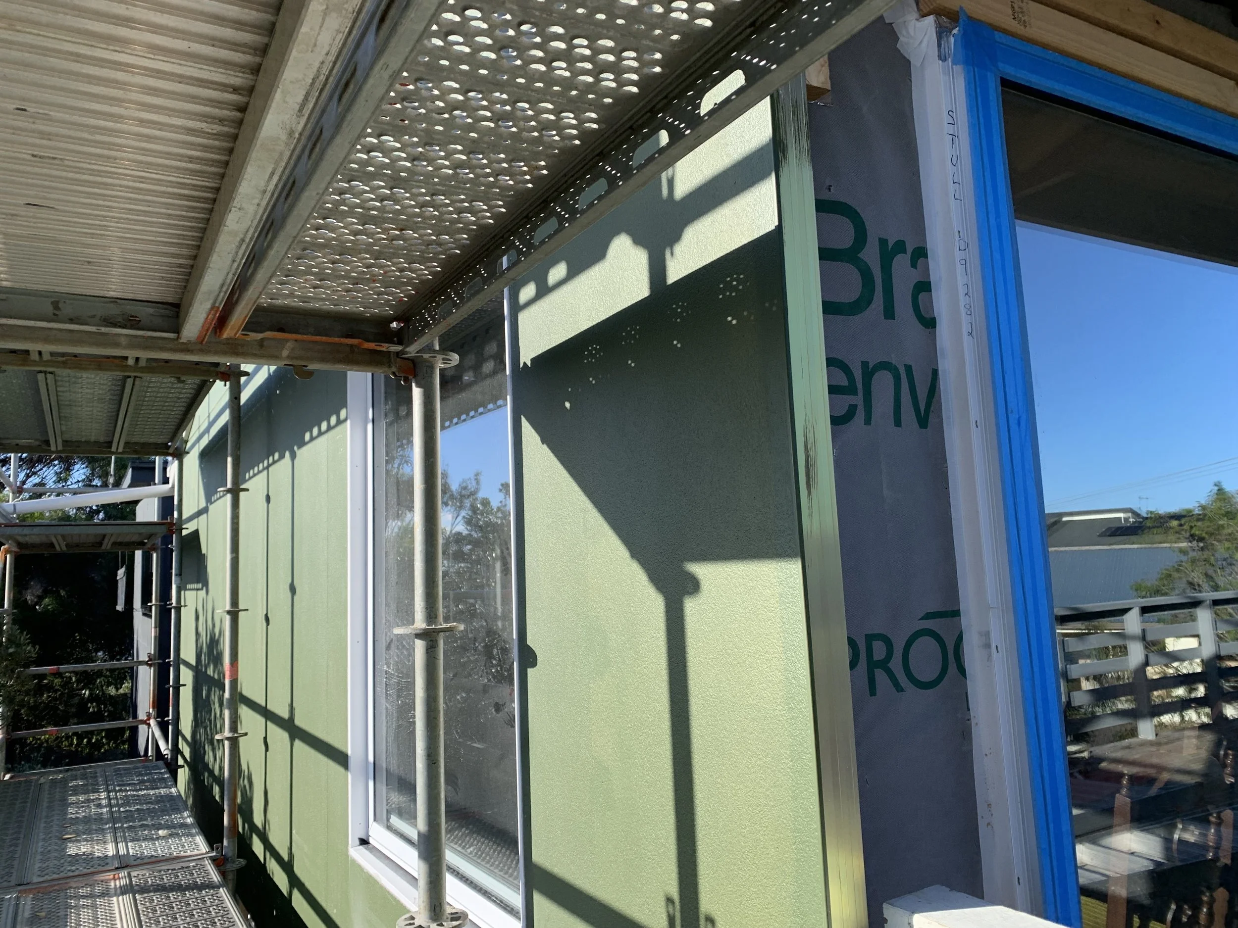 Construction site with metal scaffolding surrounding a partially built house, which has a green exterior wall, a window, and some blue and white framing.
