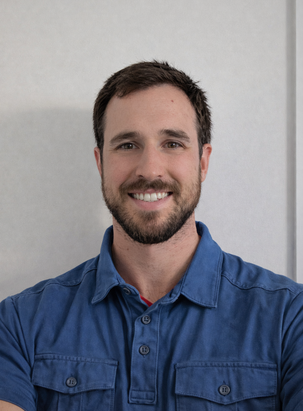 Portrait of a young man with brown hair, beard, and blue shirt, smiling against a plain light gray background.
