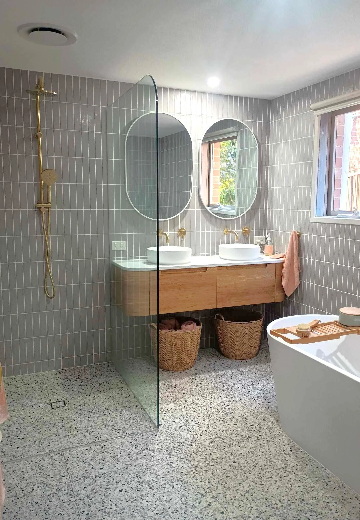 Modern bathroom with terrazzo floor, gray tiled walls, a floating wooden vanity with two vessel sinks, two oval mirrors, gold fixtures, a walk-in shower with a glass divider, a bathtub with a wooden tray, wicker baskets, and natural light from a window.