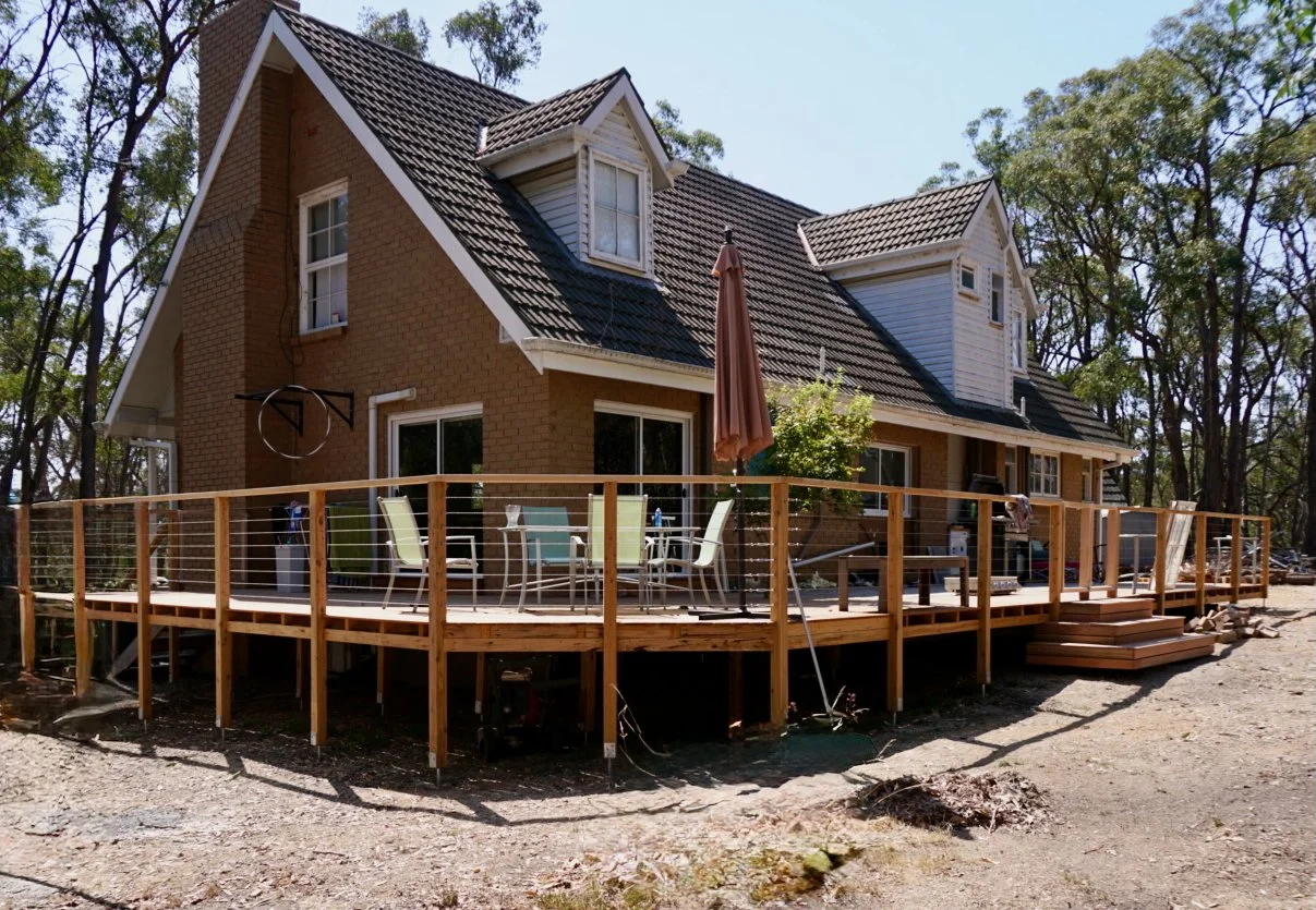 A house with a brown brick exterior, multiple gabled roofs, and a large elevated wooden deck with outdoor furniture.