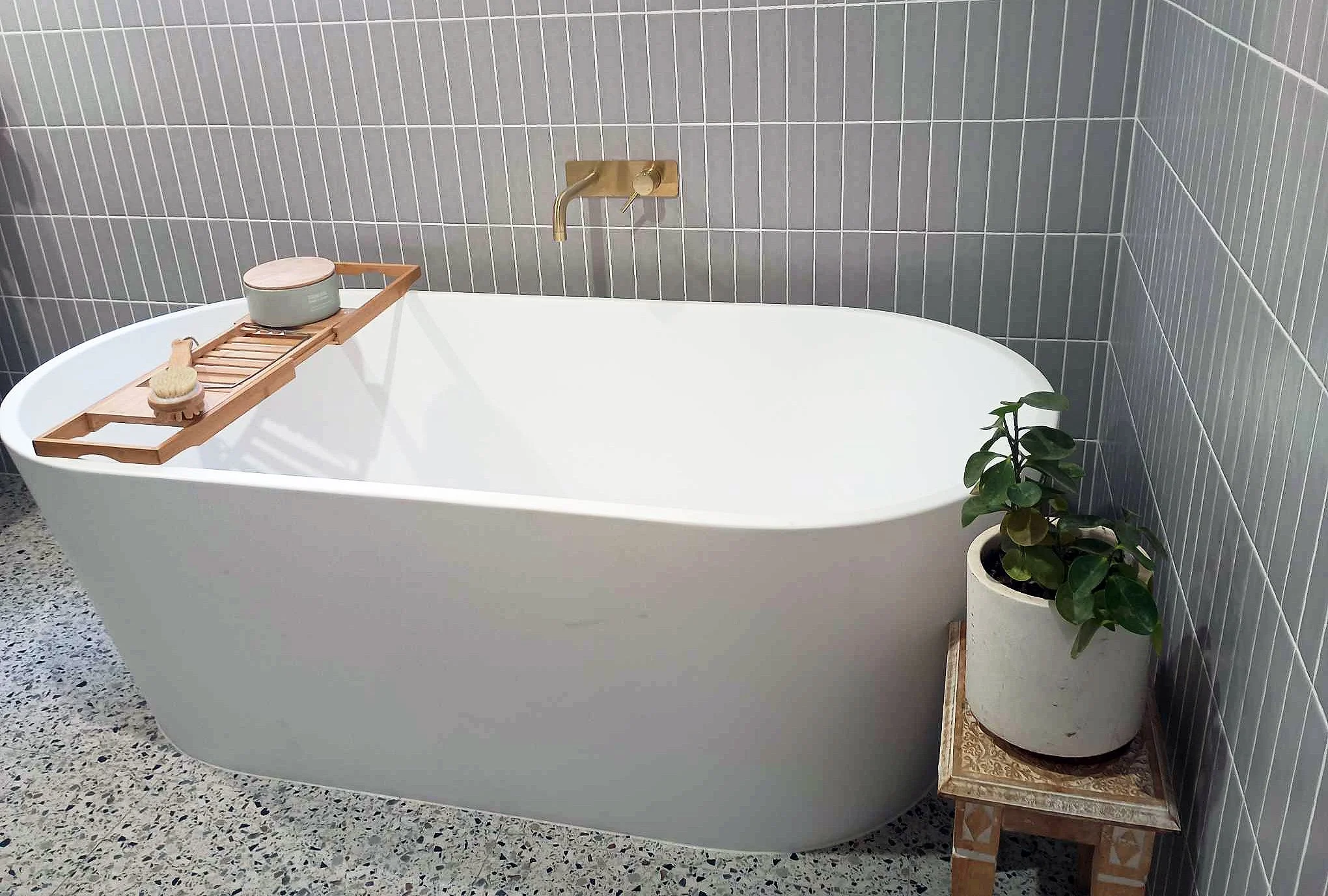 Modern bathroom with a white freestanding bathtub, gray tiled wall, gold wall-mounted faucet, wooden bath tray holding a small container and bath brush, and potted green plant on a decorative wooden table.