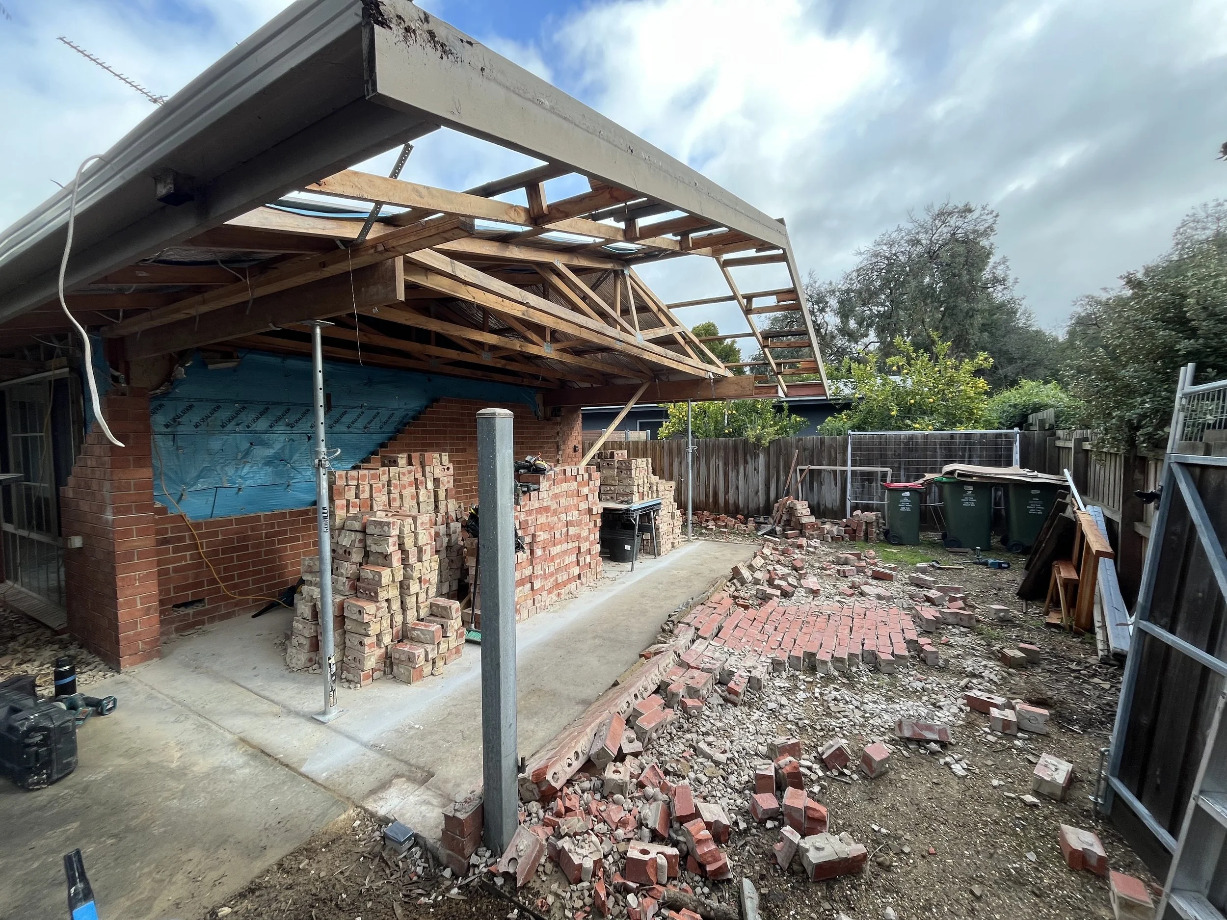 Construction site of a brick building with an ongoing roof renovation, showing piles of bricks, construction tools, and a partially completed brick wall.